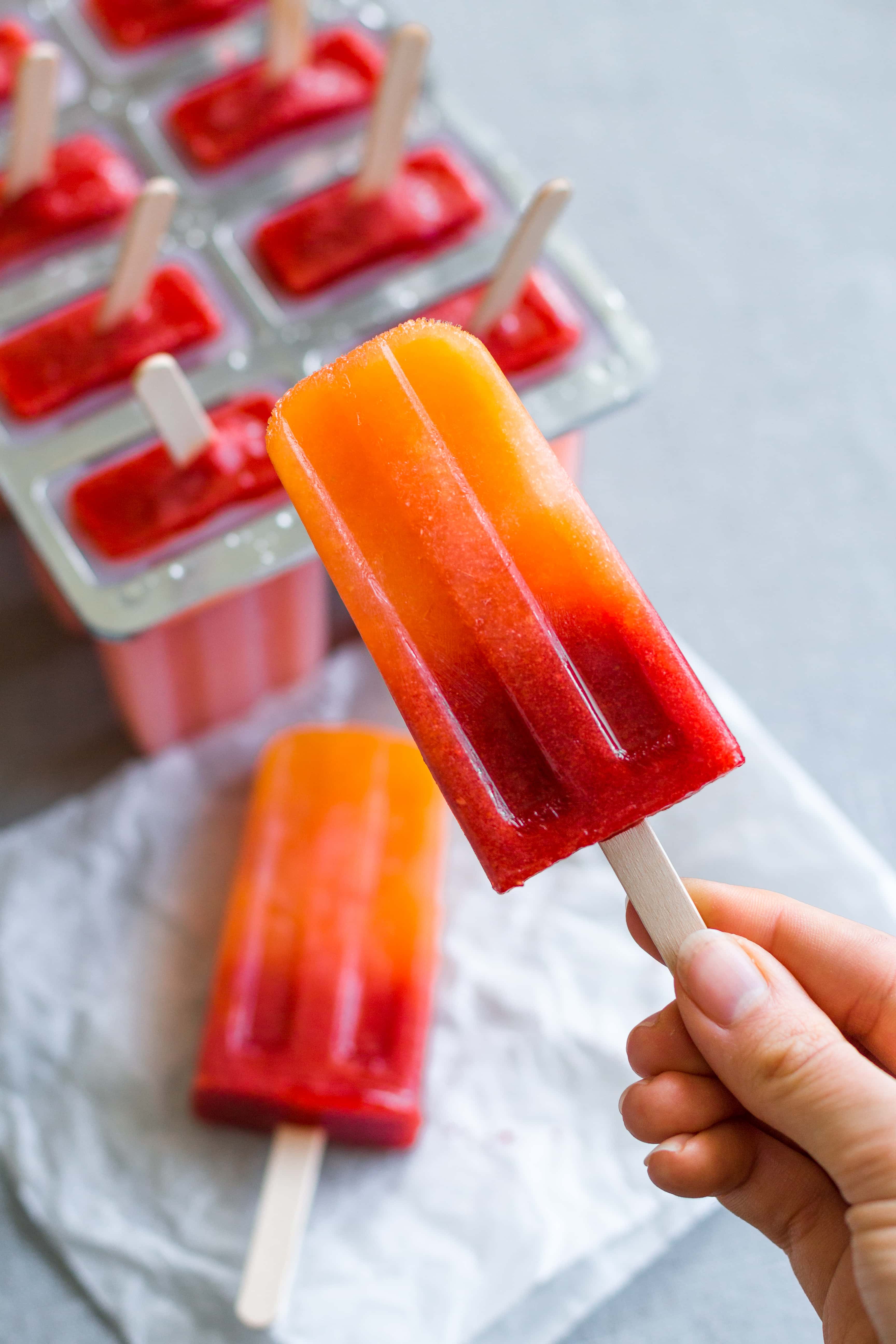 A hand holding an ombre orange and red popsicle. More popsicles blurry in the background, one on paper lying on the table and a few in a popsicle mold.