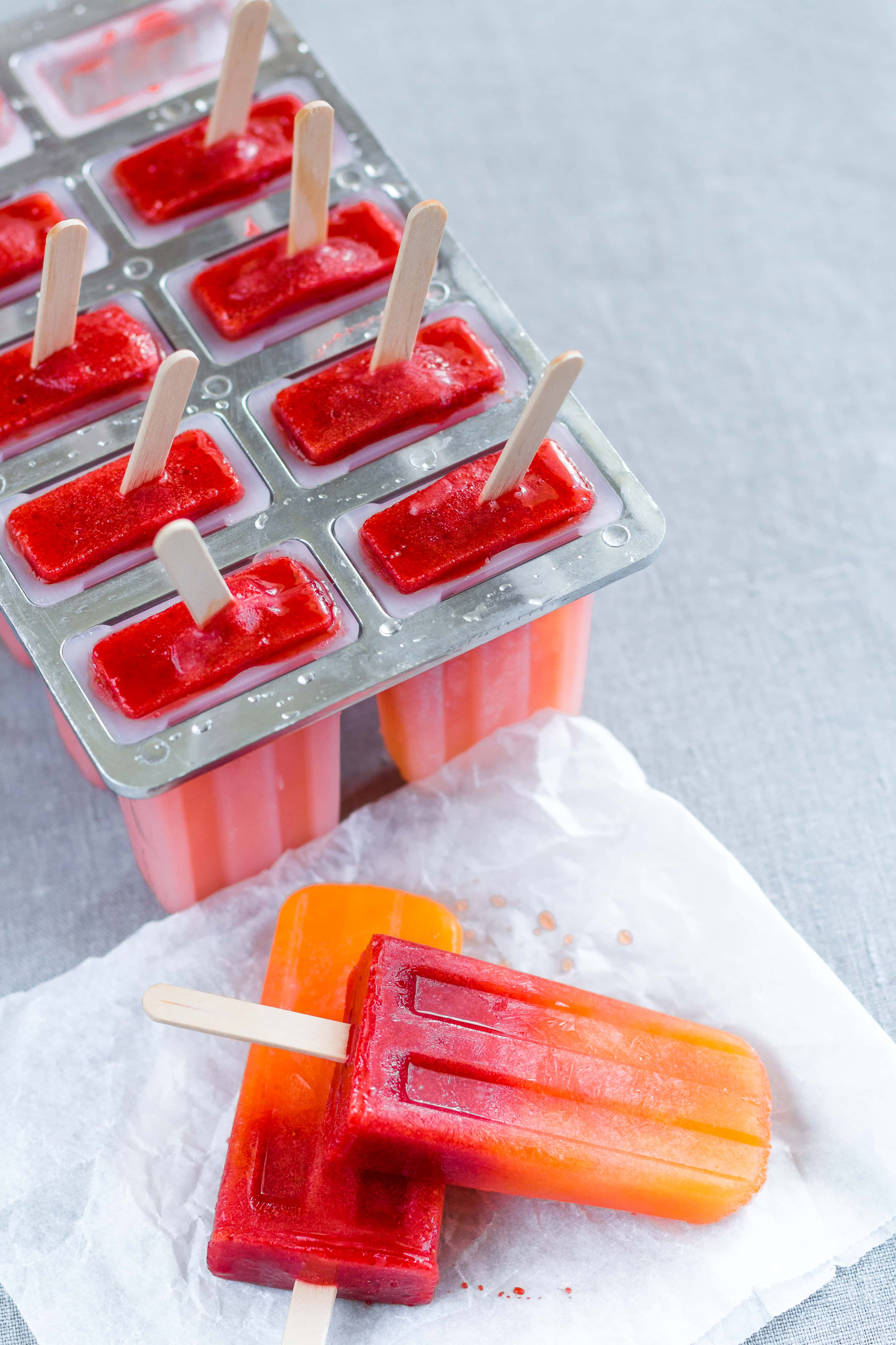 Orange Strawberry Popsicles in the popsicle molds with popsicle sticks sticking out of the ice cream. On a grey table cloth.