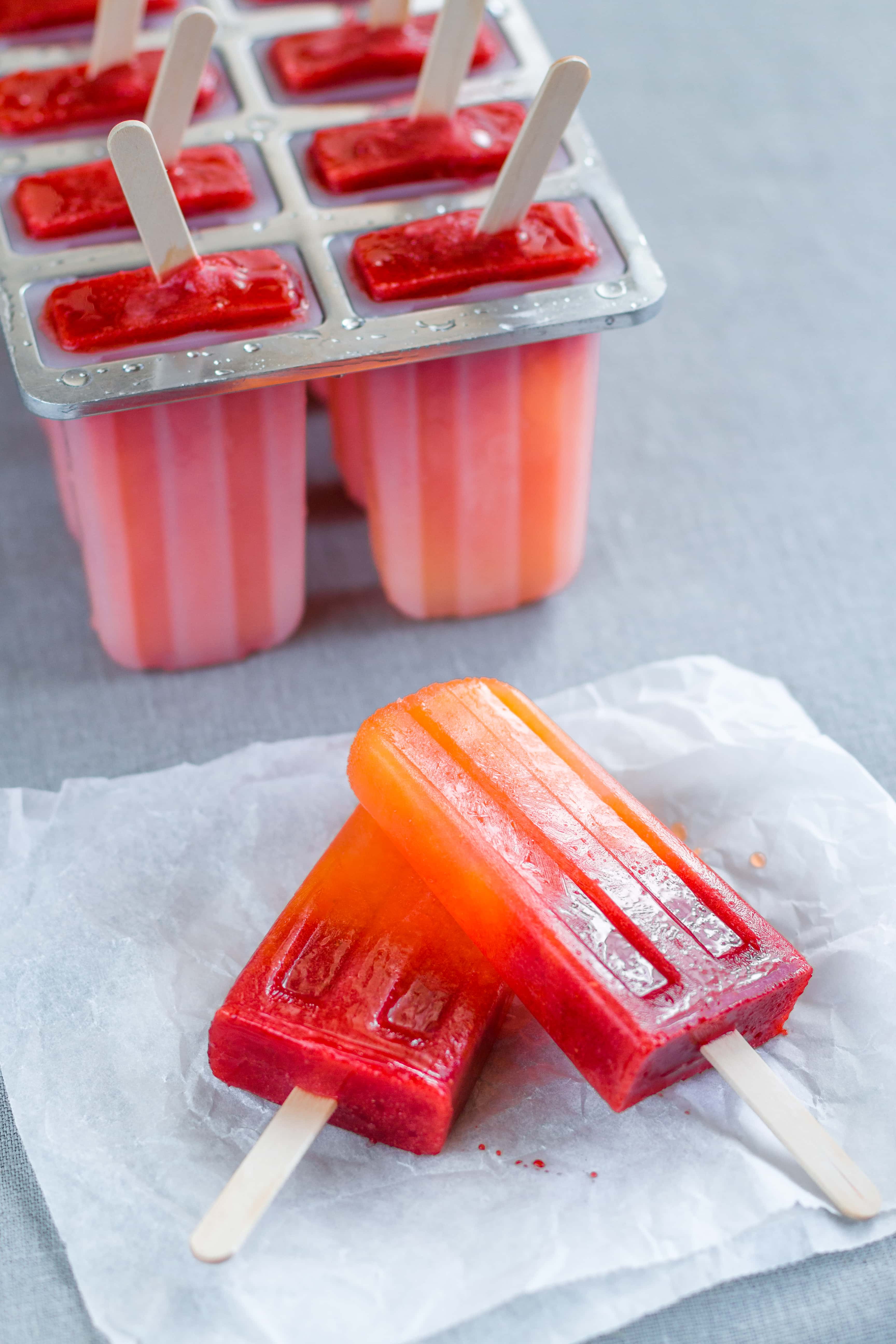Orange Strawberry Sunrise Popsicles on grey table cloth. Six popsicles still in the popsicle molds on the top left coming out of the frame. In the middle, there are two popsicles lying on some parchment paper in front of the popsicles in the molds.