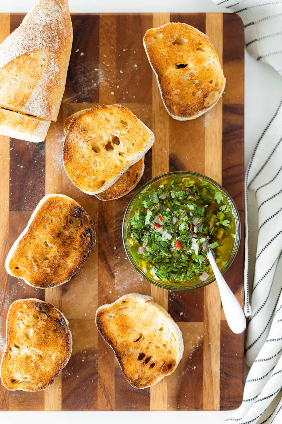 Sliced toasted baguette on a wooden cutting board, with chimichurri sauce in a glass bowl