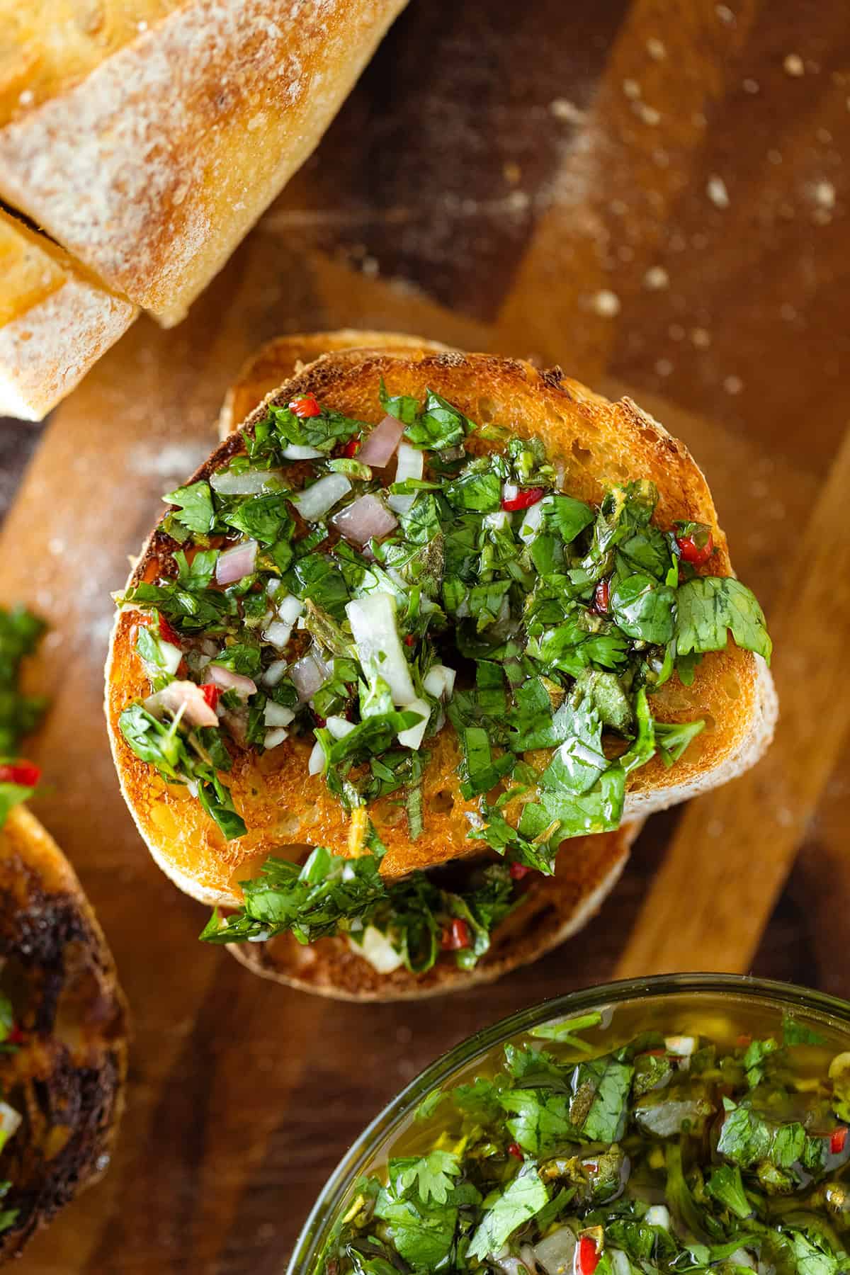 A close up of a slice of toasted baguette with chimichurri sauce on a cutting board