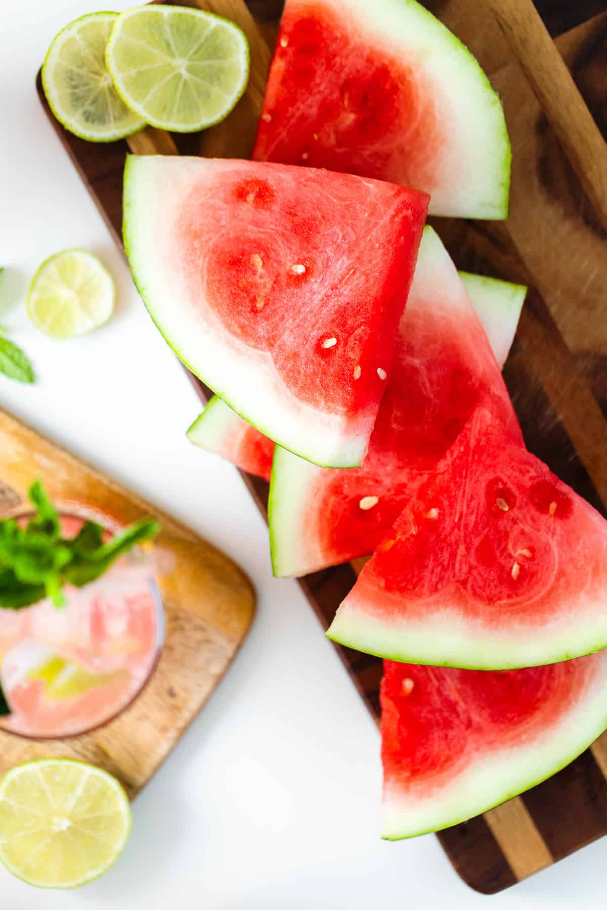 Sliced watermelon on a dark wood cutting board with slices of lime and a watermelon coconut rum cooler in the left bottom corner