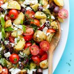 Greek Potato Salad with Feta, Olives, and fresh herbs on an oval platter - close up. Bright blue background