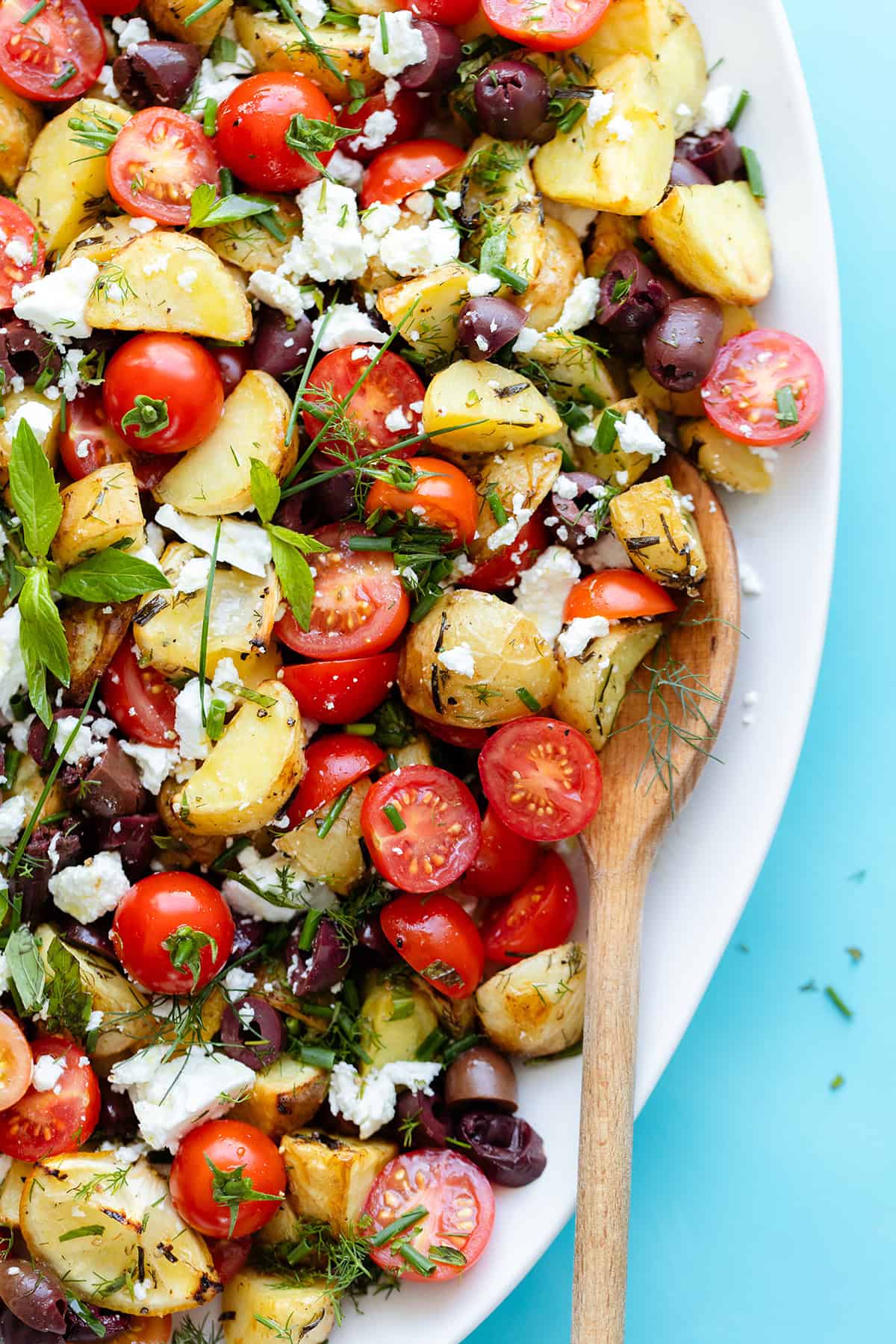 Greek Potato Salad with Feta, Olives, and fresh herbs on an oval platter - close up. Bright blue background