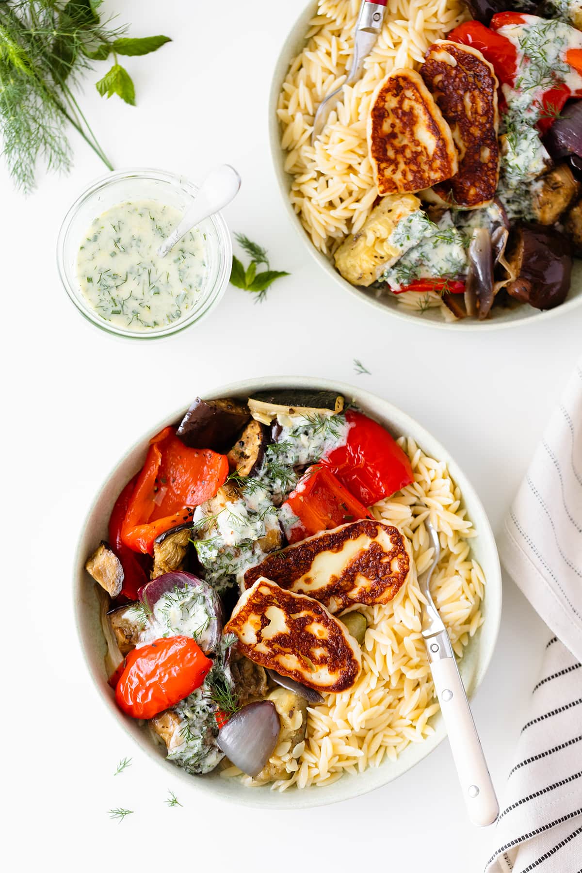 Halloumi Orzo Bowl with Roasted Vegetables and Yogurt Dill Sauce on two plates on a white table decorated with fresh herbs and a striped cloth on the right side of the frame. Overhead shot