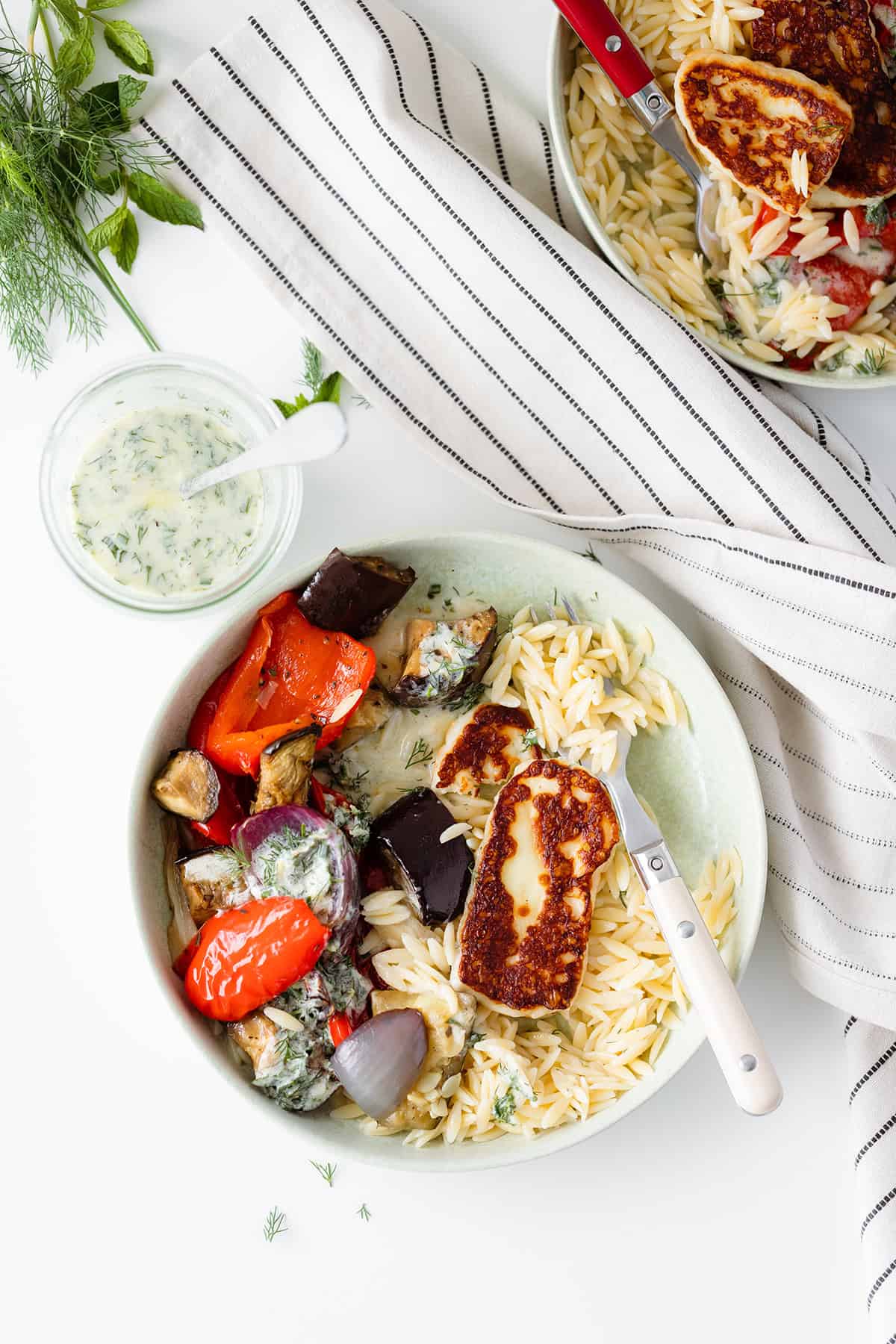 Halloumi Orzo Bowl with Roasted Vegetables and Yogurt Dill Sauce on two plates on a white table decorated with fresh herbs and a striped cloth on the right side of the frame. Overhead shot