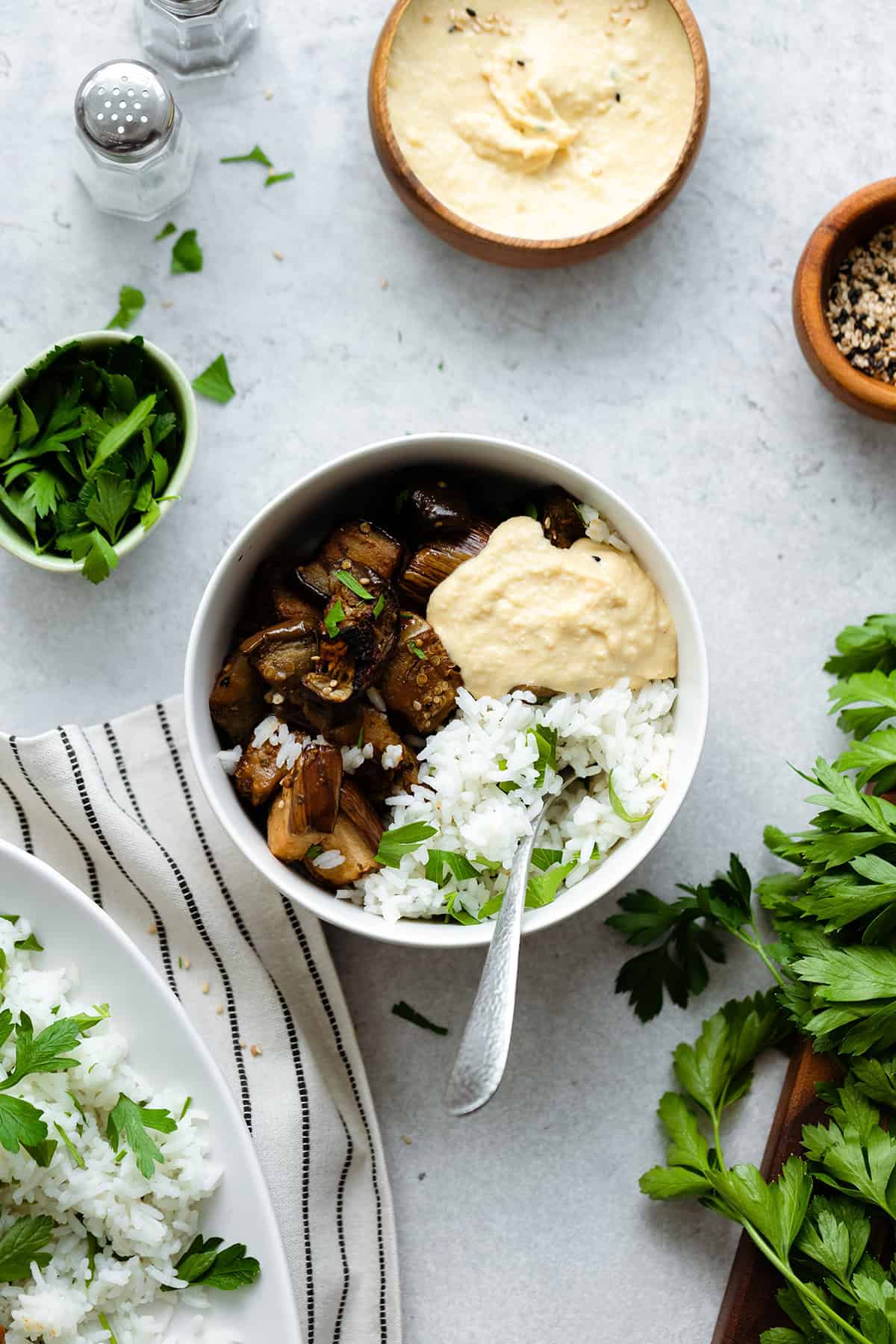 Spicy Garlic Miso Roasted Eggplant in a small bowl with white rice, parsley, and hummus. Grey background