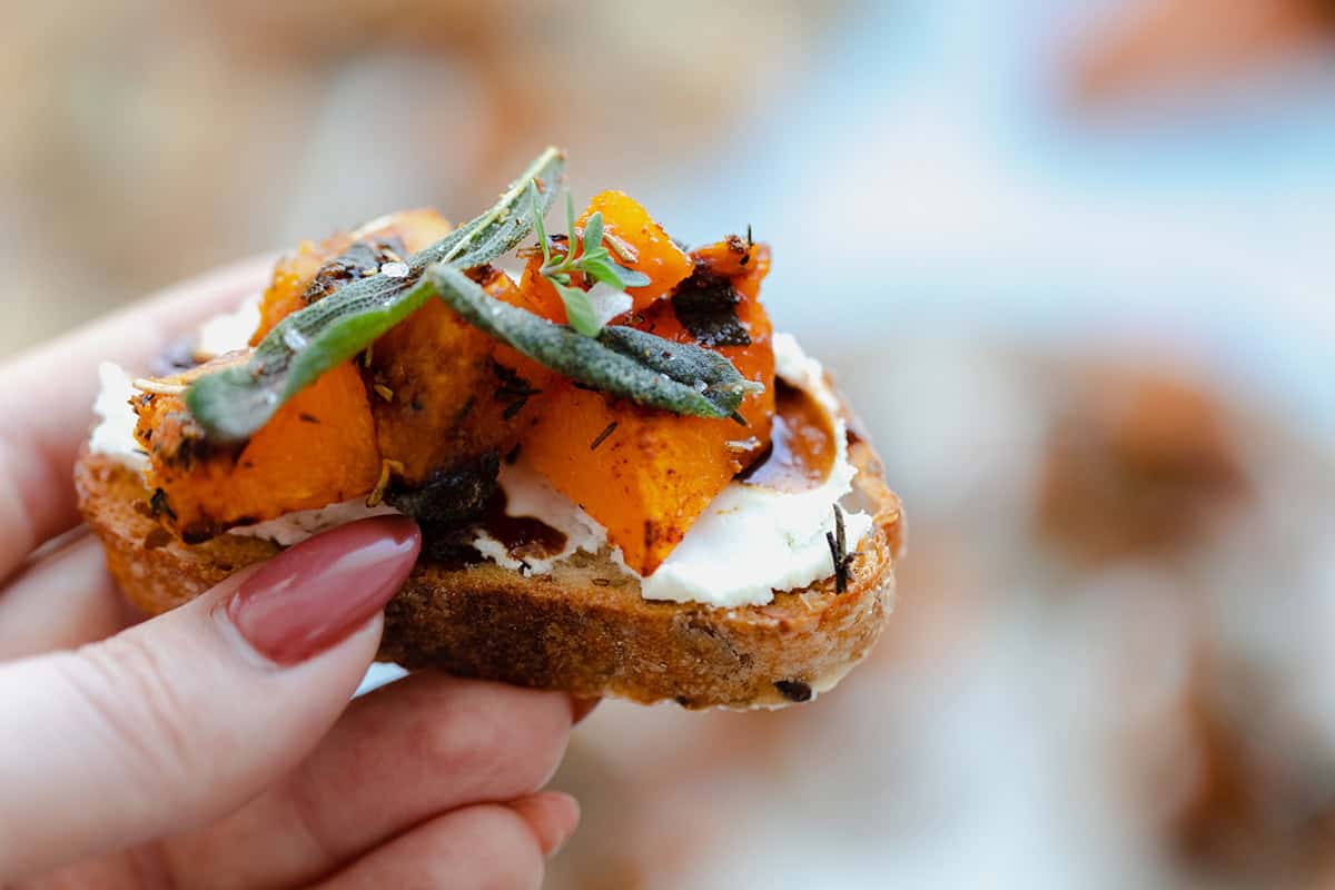 A photo of a hand holding Butternut Squash Crostini with Goat Cheese and Fig Balsamic Glaze