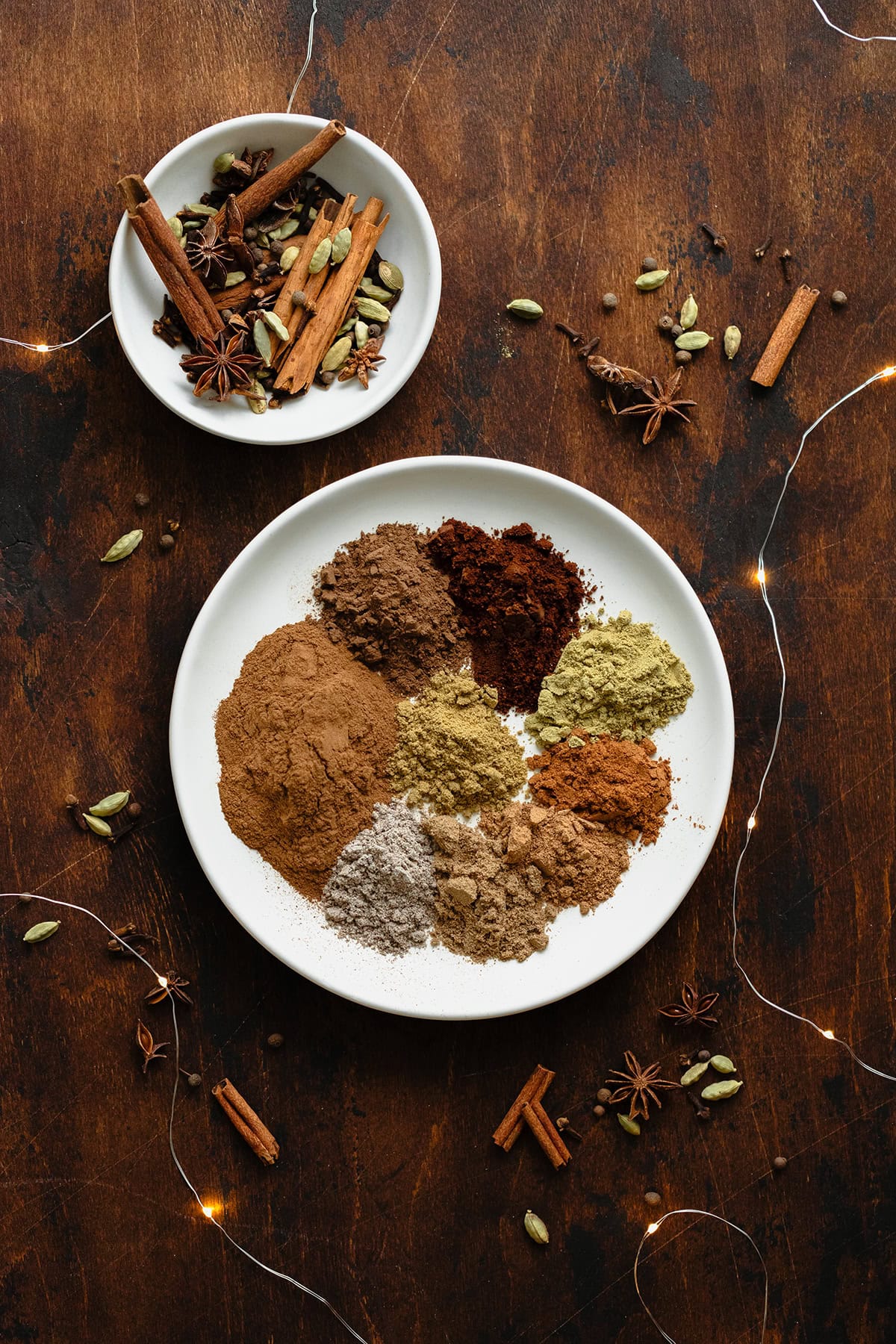 A photo of all the spices used in the Christmas Spice Blend on a white plate. On a dark wooden background and fairy lights.