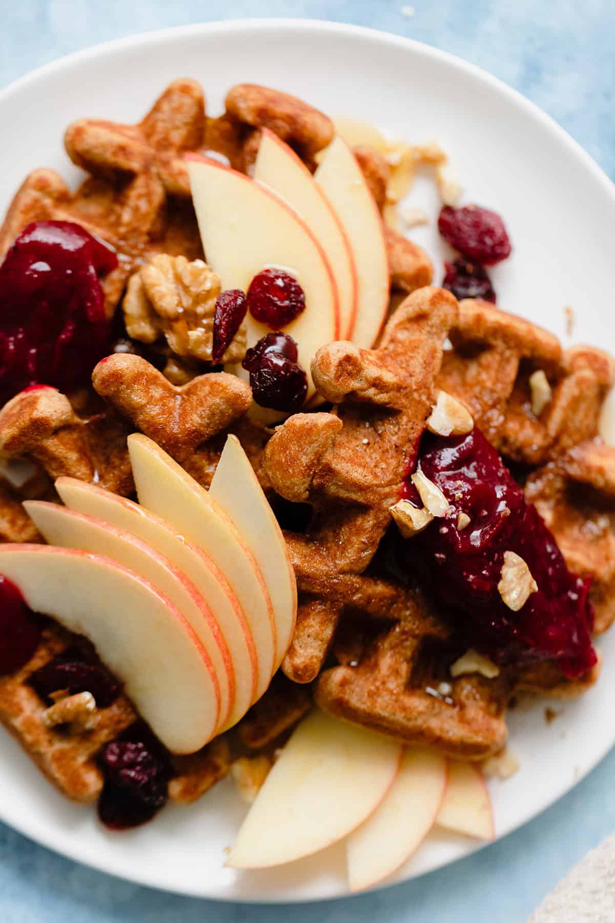 An overhead photo fo Gluten-Free Apple Butter Waffles with apples, cranberries, and walnuts on blue background.