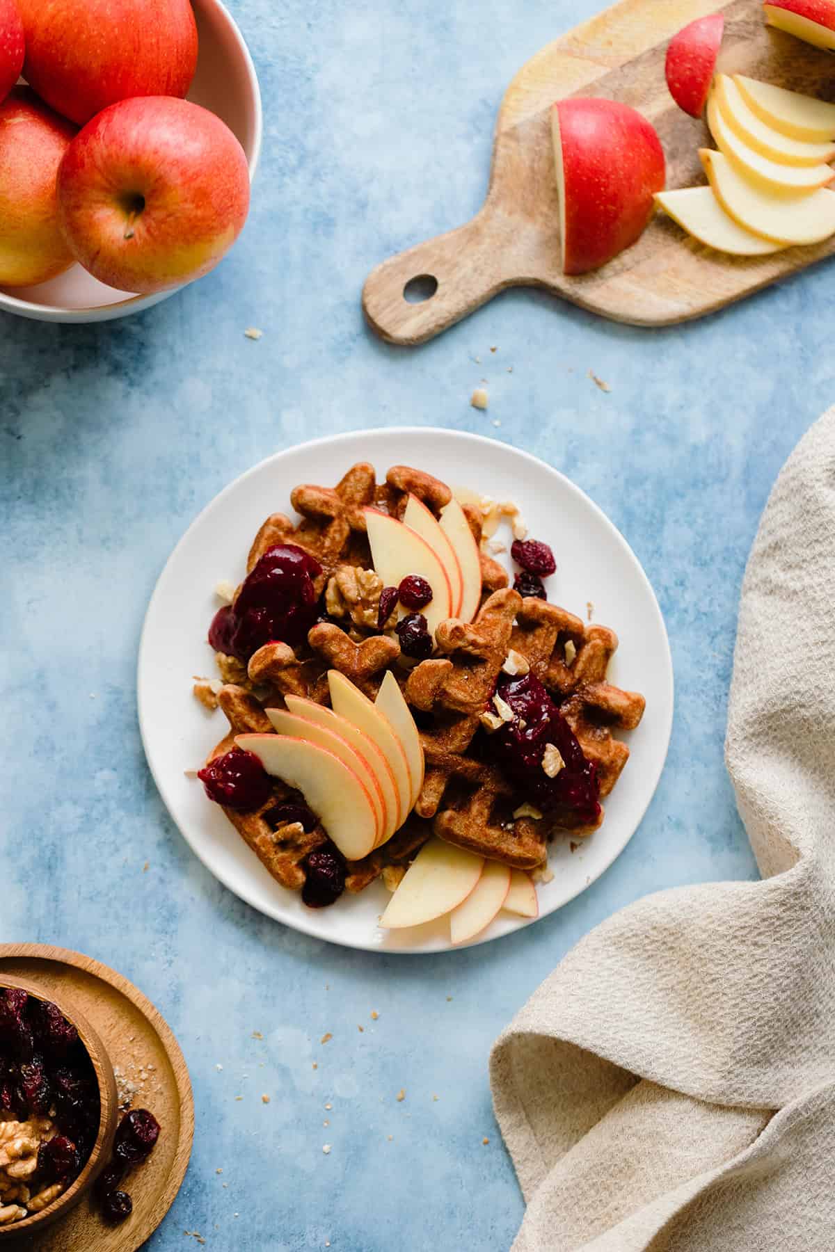 An overhead photo fo Gluten-Free Apple Butter Waffles with apples, cranberries, and walnuts on blue background.