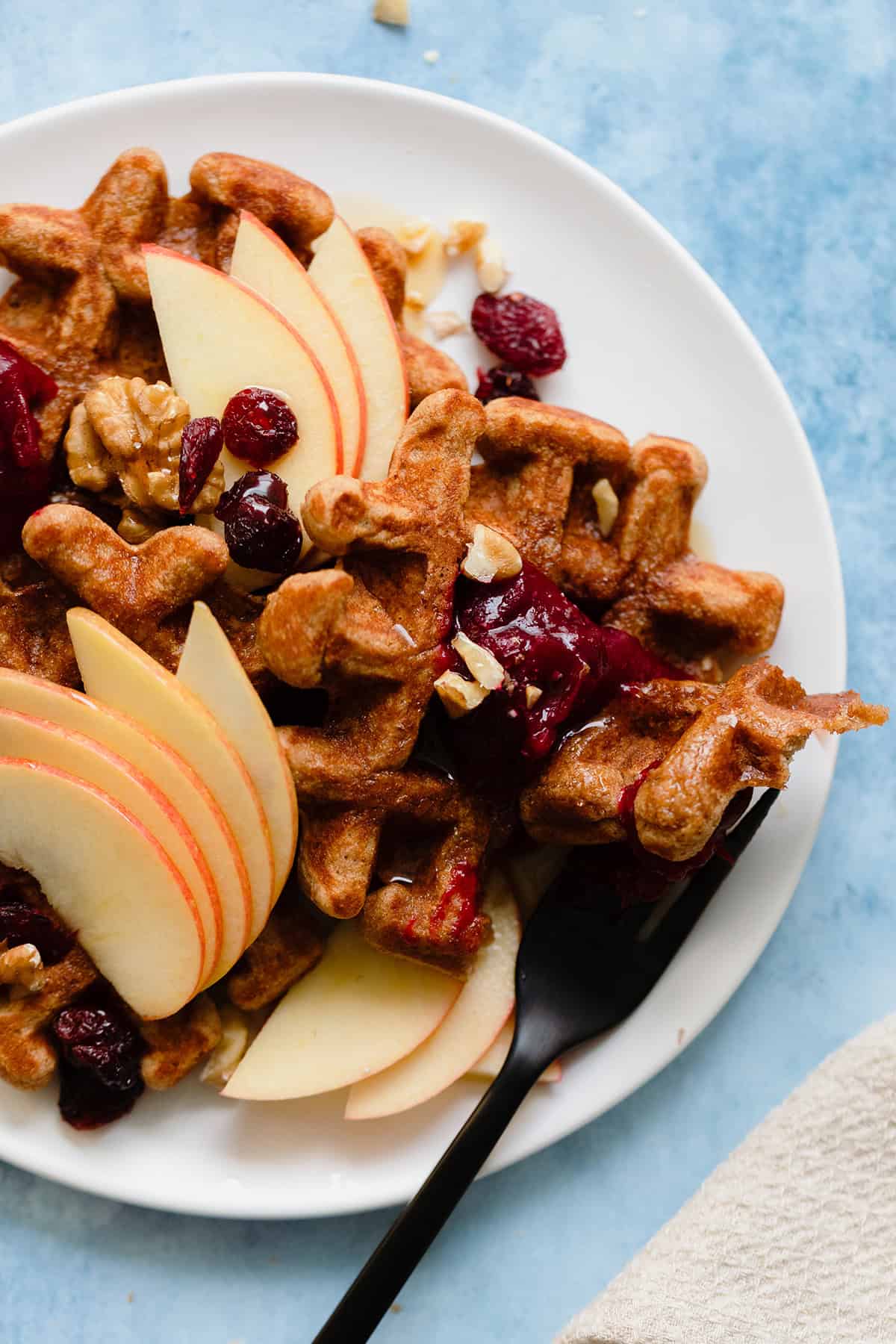 An overhead photo fo Gluten-Free Apple Butter Waffles with apples, cranberries, and walnuts on blue background.