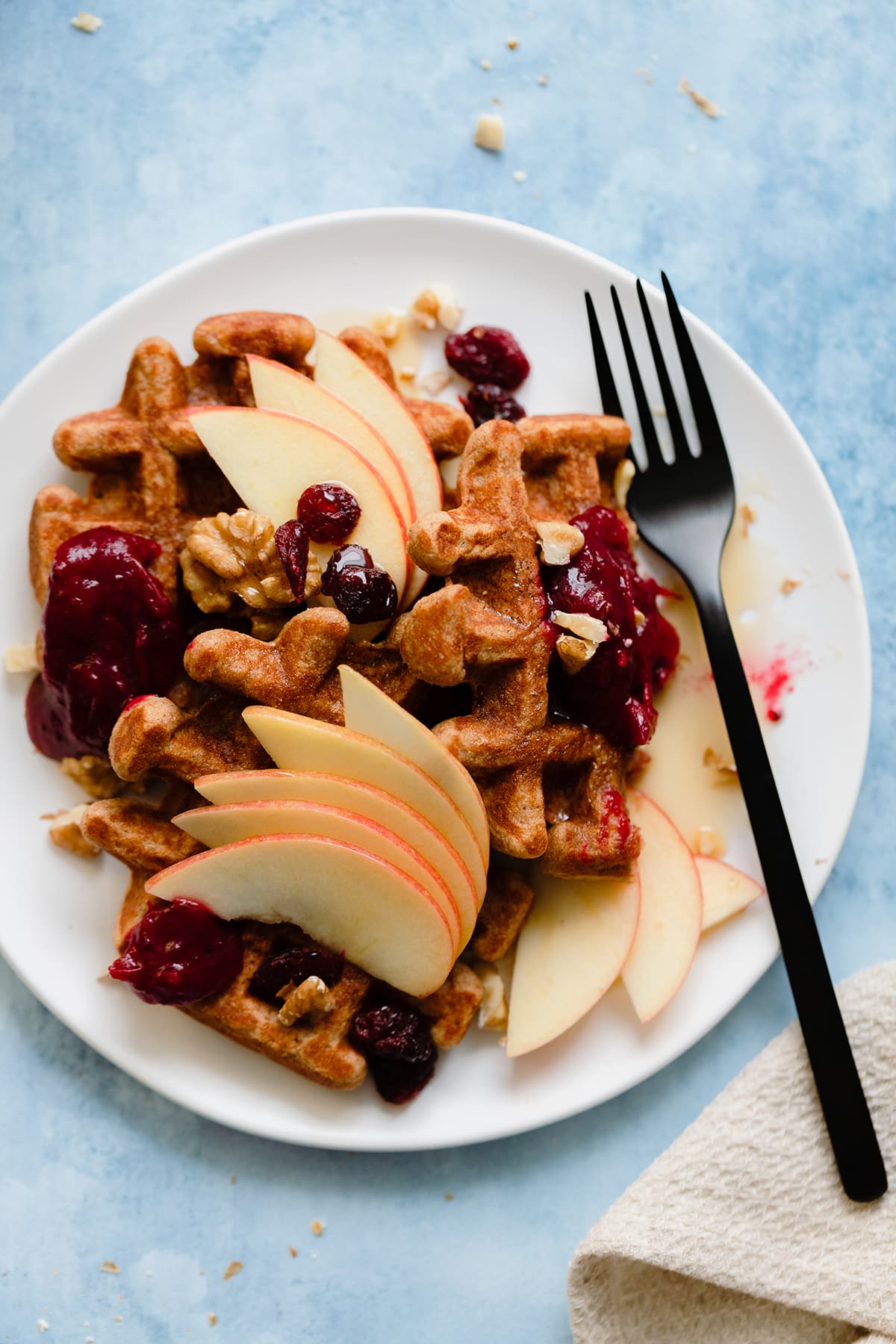 An overhead photo fo Gluten-Free Apple Butter Waffles with apples, cranberries, and walnuts on blue background.