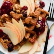 An overhead photo fo Gluten-Free Apple Butter Waffles with apples, cranberries, and walnuts on blue background.