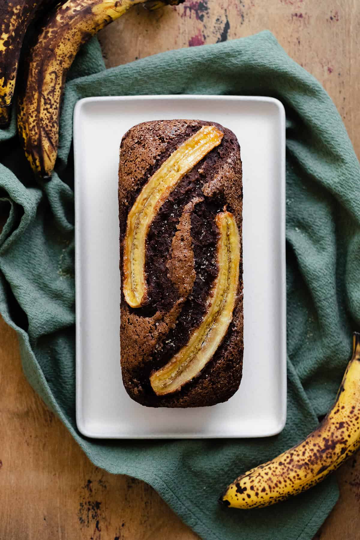 A photo of Caramelized Chocolate Banana Bread on a beige plate, green kitchen towel, and light wooden table.