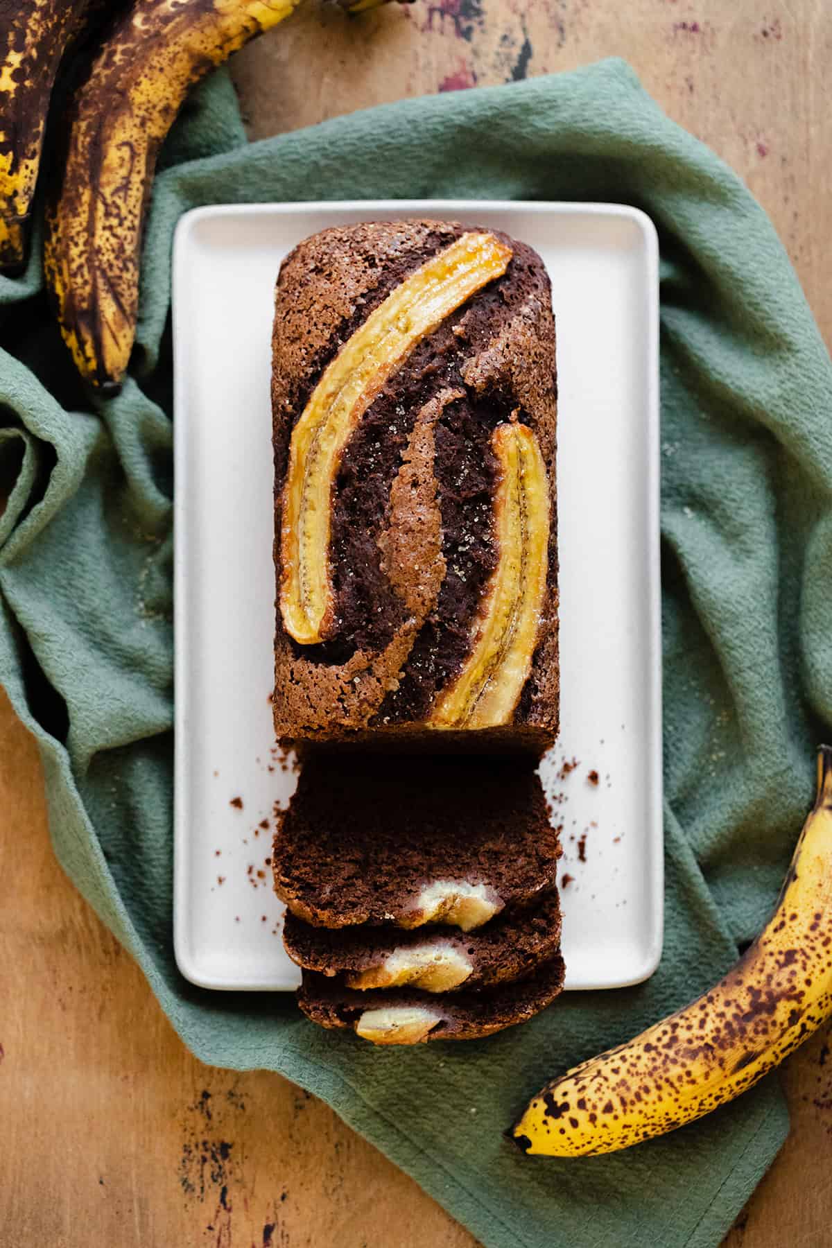 A photo of Caramelized Chocolate Banana Bread on a beige plate, green kitchen towel, and light wooden table.