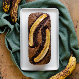A photo of Caramelized Chocolate Banana Bread on a beige plate, green kitchen towel, and light wooden table.