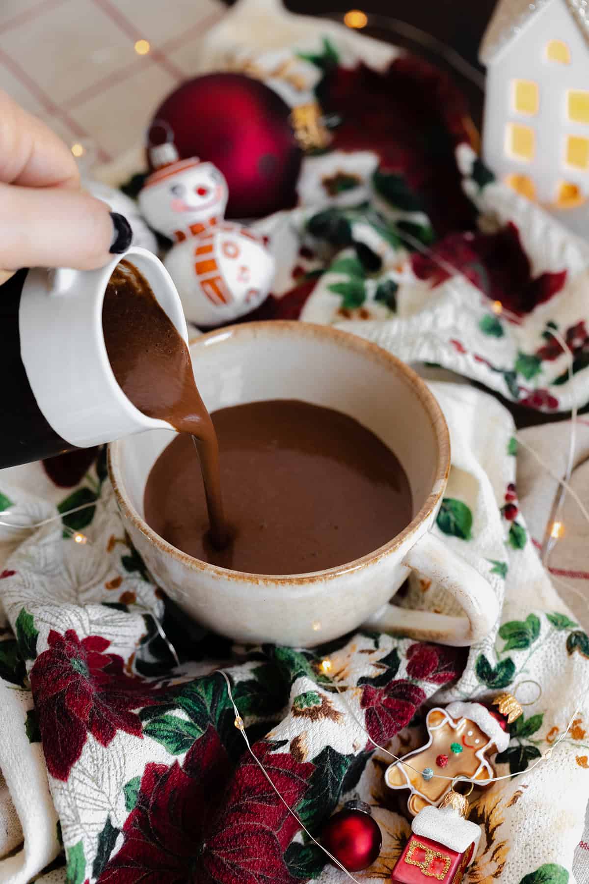 A shot of the hot chocolate being poured into a cup on a Christmas themed tablecloth.