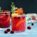 Cranberry Orange Margarita in a glass with a gold rim on a blue table with a black background.