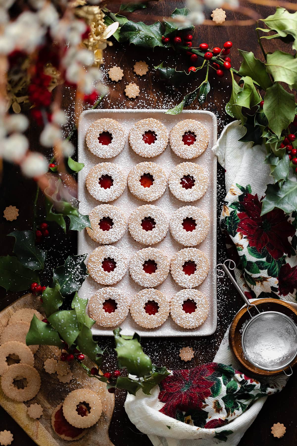 Gluten-Free Linzer Cookies with raspberry jam and a dusting of powdered sugar on a beige plate. Decorated with Christmas decorations all around the plate