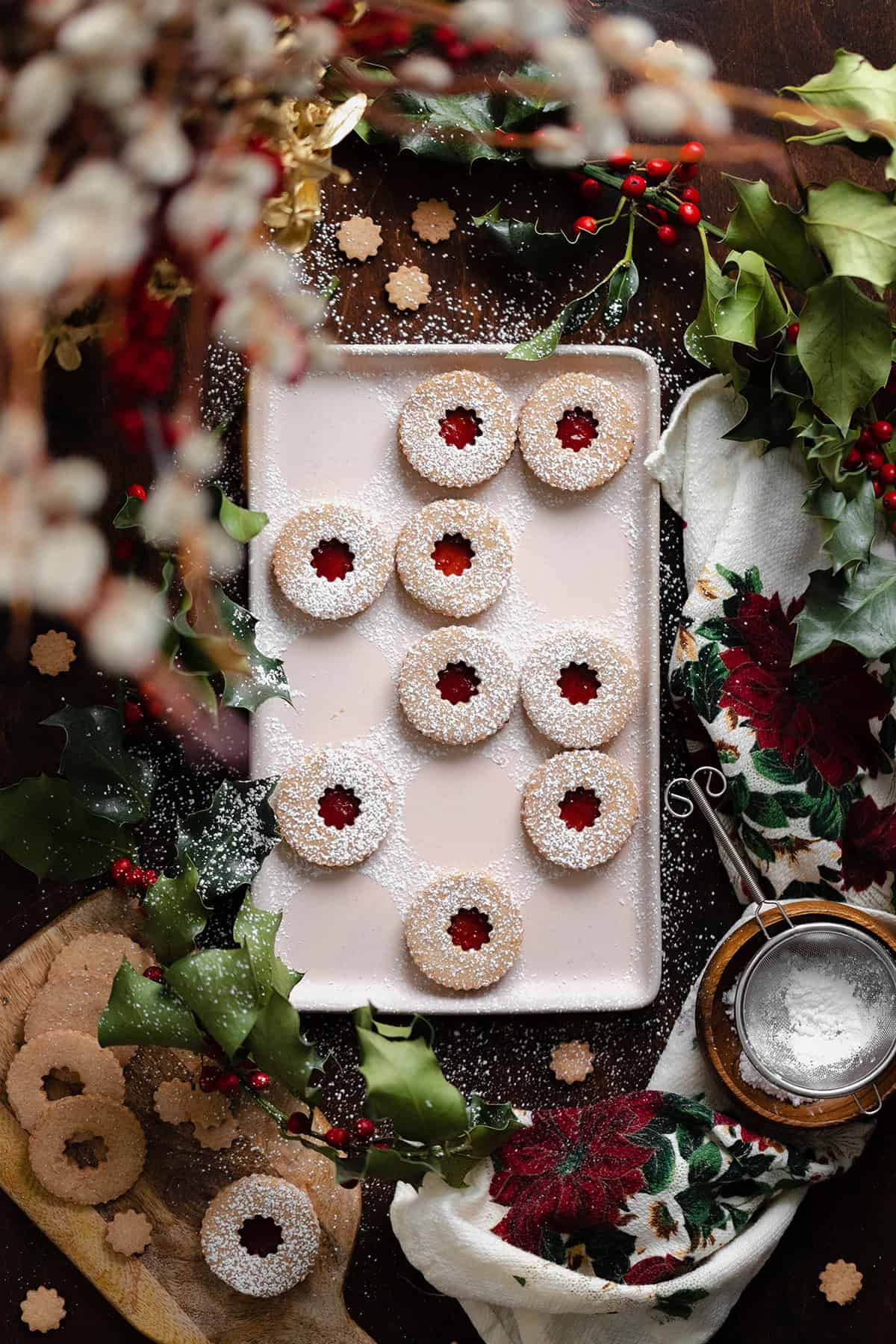 Gluten-Free Linzer Cookies with raspberry jam and a dusting of powdered sugar on a beige plate. Decorated with Christmas decorations all around the plate