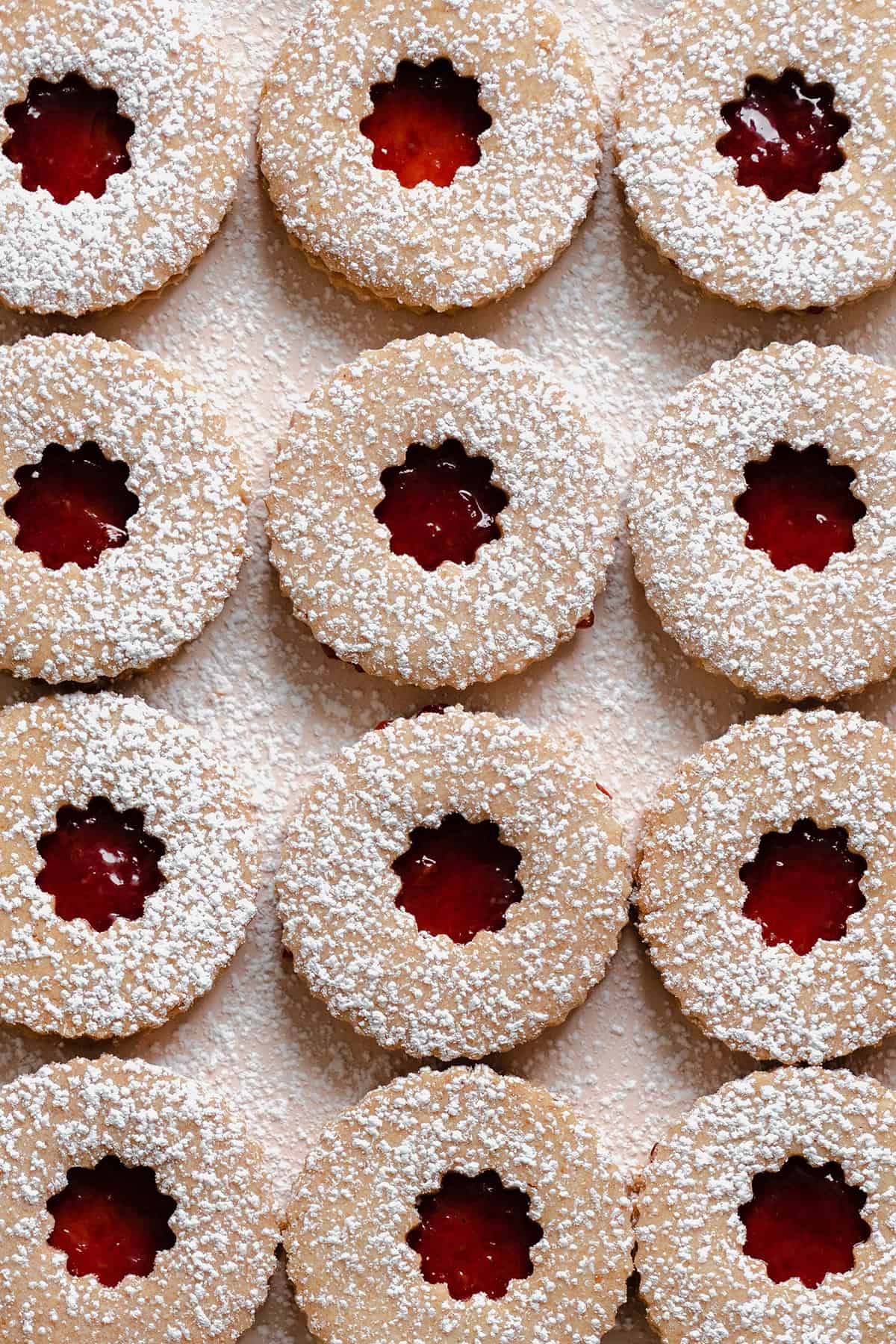 A close up image of Linzer Cookies with raspberry jam and a dusting of powdered sugar on a beige plate.