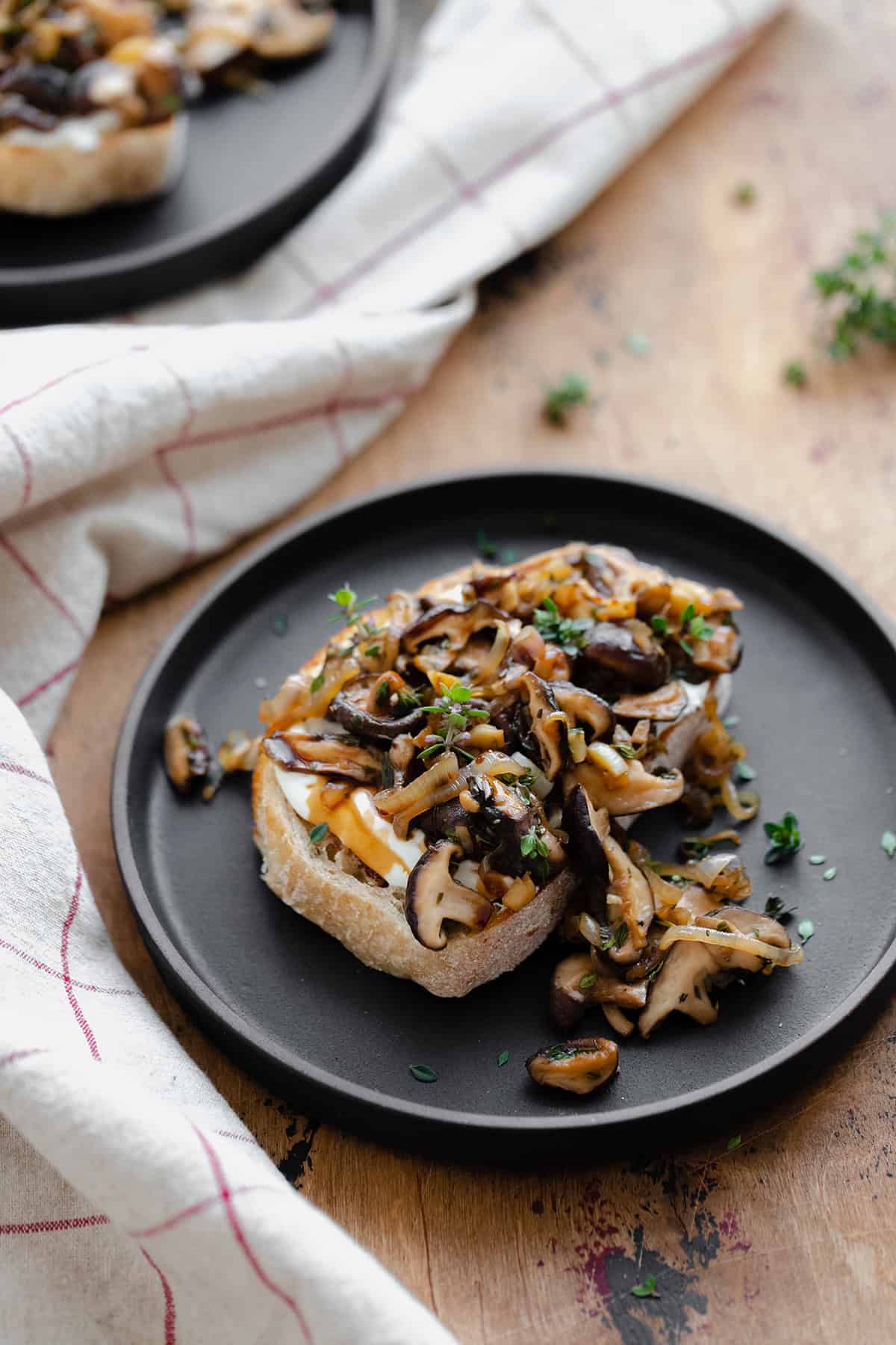 Whipped Goat Cheese Toast with Shiitake Mushroom on a black plate and a light wooden table. Natural cotton napkin on the left side.