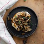 Whipped Goat Cheese Toast with Shiitake Mushroom on a black plate and a light wooden table. Natural cotton napkin on the left side.