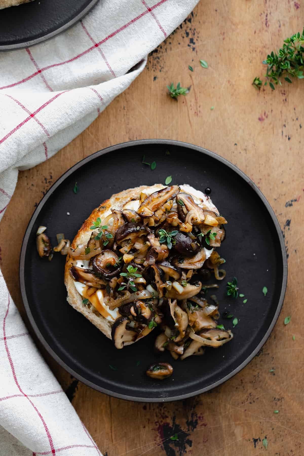 Whipped Goat Cheese Toast with Shiitake Mushroom on a black plate and a light wooden table. Natural cotton napkin on the left side.