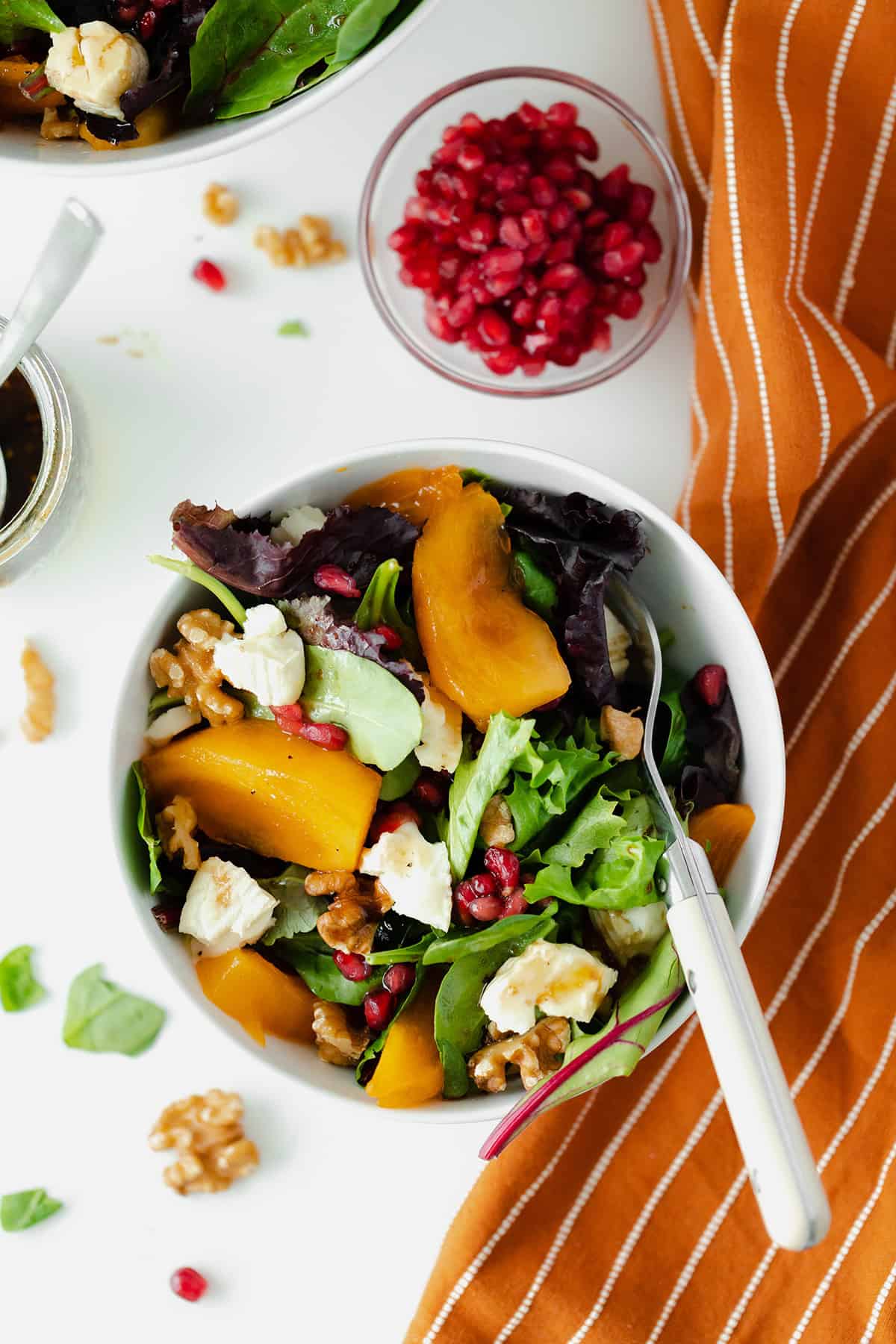 Persimmon Salad with Goat Brie and Pomegranate seeds in a white bowl on a white background with an orange napkin on the right side