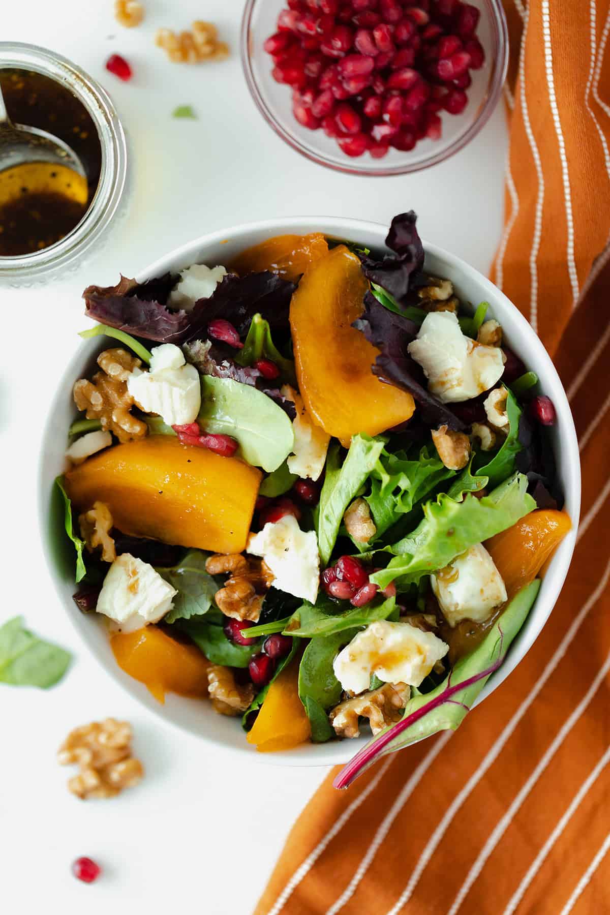 Persimmon Salad with Goat Brie and Pomegranate seeds in a white bowl on a white background with an orange napkin on the right side