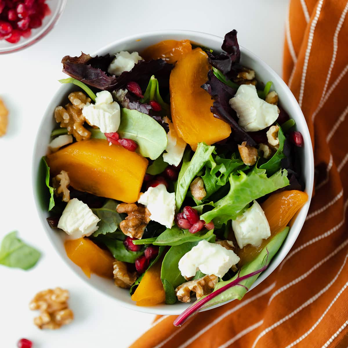 Persimmon Salad with Goat Brie and Pomegranate seeds in a white bowl on a white background with an orange napkin on the right side
