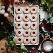 Gluten-Free Linzer Cookies with raspberry jam and a dusting of powdered sugar on a beige plate. Decorated with Christmas decorations all around the plate