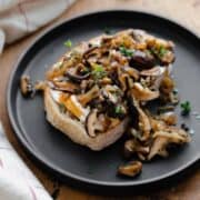 Whipped Goat Cheese Toast with Shiitake Mushroom on a black plate and a light wooden table. Natural cotton napkin on the left side.