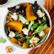 Persimmon Salad with Goat Brie and Pomegranate seeds in a white bowl on a white background with an orange napkin on the right side