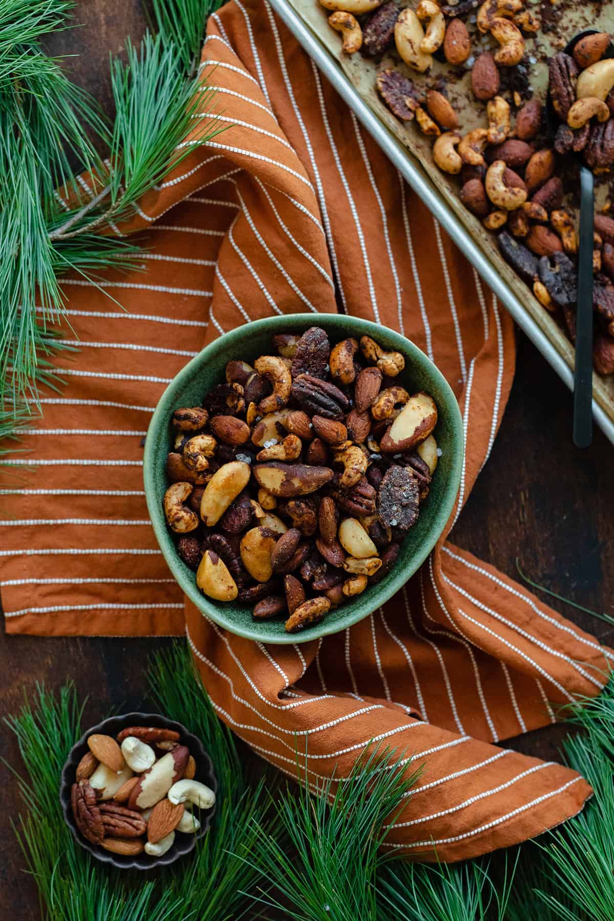 Mixed roasted nuts in a green bowl on an orange and white striped napkin. Pine needles peeking into the shot in the top left, bottom left, and bottom right corners. Baking sheet with more nuts in the top right corner.