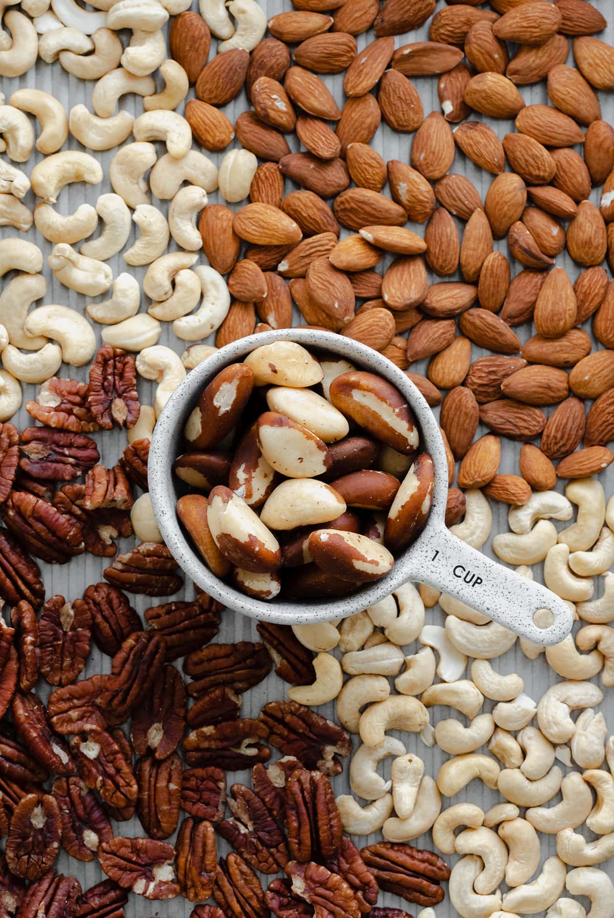 Raw nuts spread on a baking sheet. Divided in four kinds. Top left are cashews, top right almonds, bottom left pecans, bottom right more cashews. In the middle there's a 1 cup measuring cup with brazil nuts.