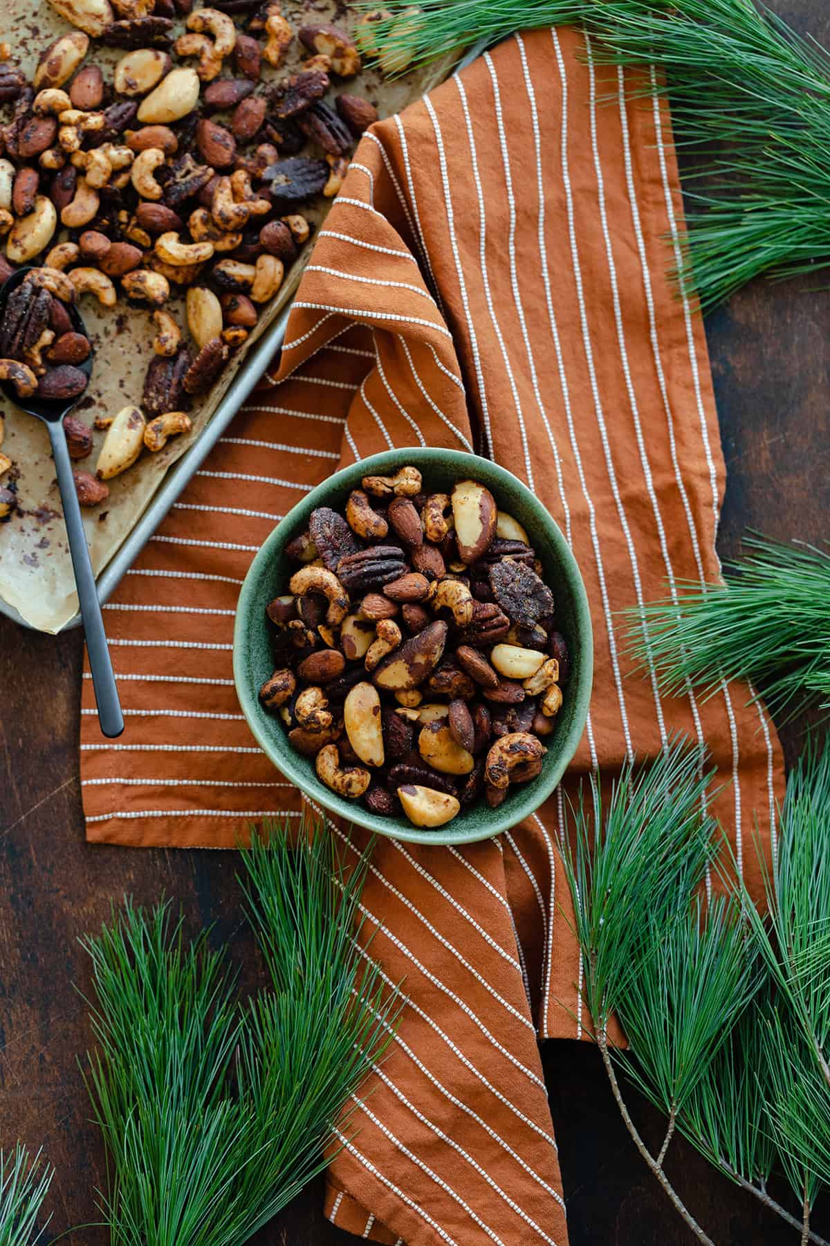 Mixed roasted nuts in a green bowl on an orange and white striped napkin. Pine needles peeking into the shot in the top right, bottom left, and bottom right corners.