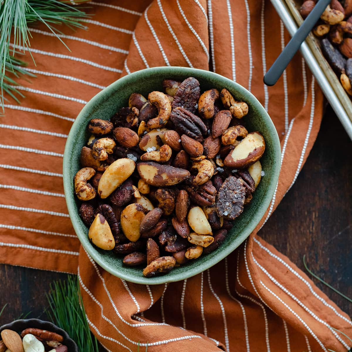 Mixed roasted nuts in a green bowl on an orange and white striped napkin. Pine needles peeking into the shot in the top left and bottom left corner.