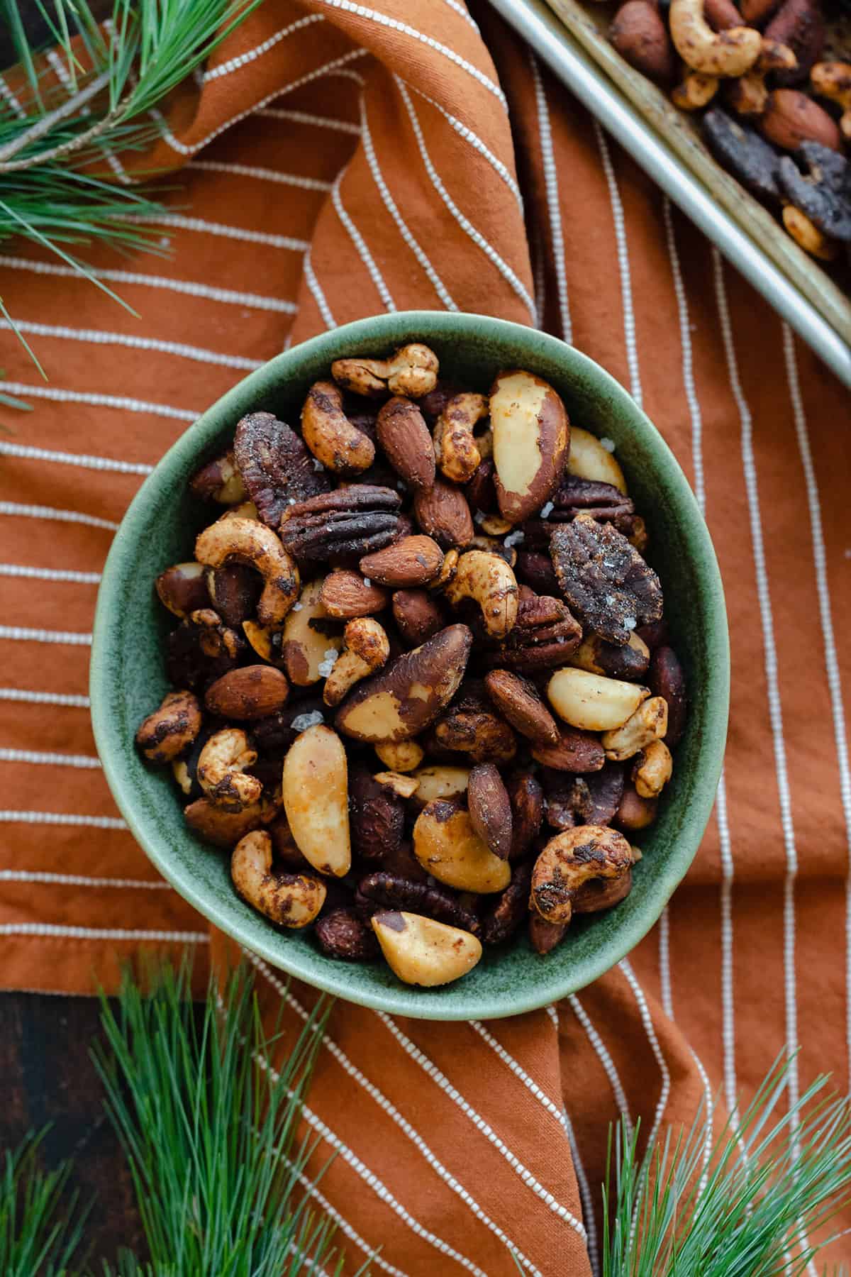 Mixed roasted nuts in a green bowl on an orange and white striped napkin. Pine needles peeking into the shot in the top left, bottom left, and bottom right corners.