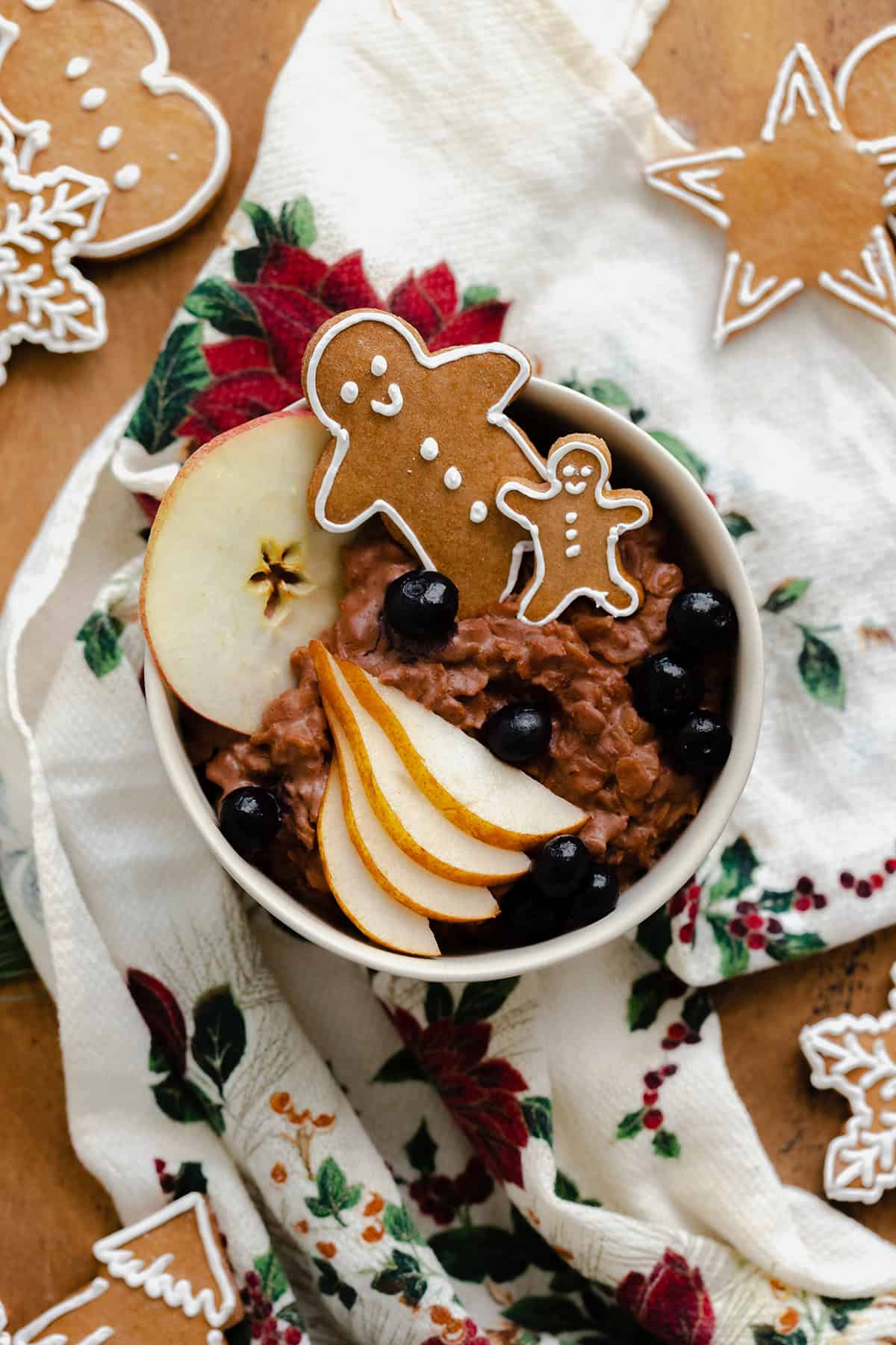 An overhead photo of Gingerbread oatmeal in a beige bowl garnished with sliced pear, a slice of apple, a few blueberries, and two gingrbread men. On light wooden background with a christmas tea towel under the bowl and gingerbread cookies around it.