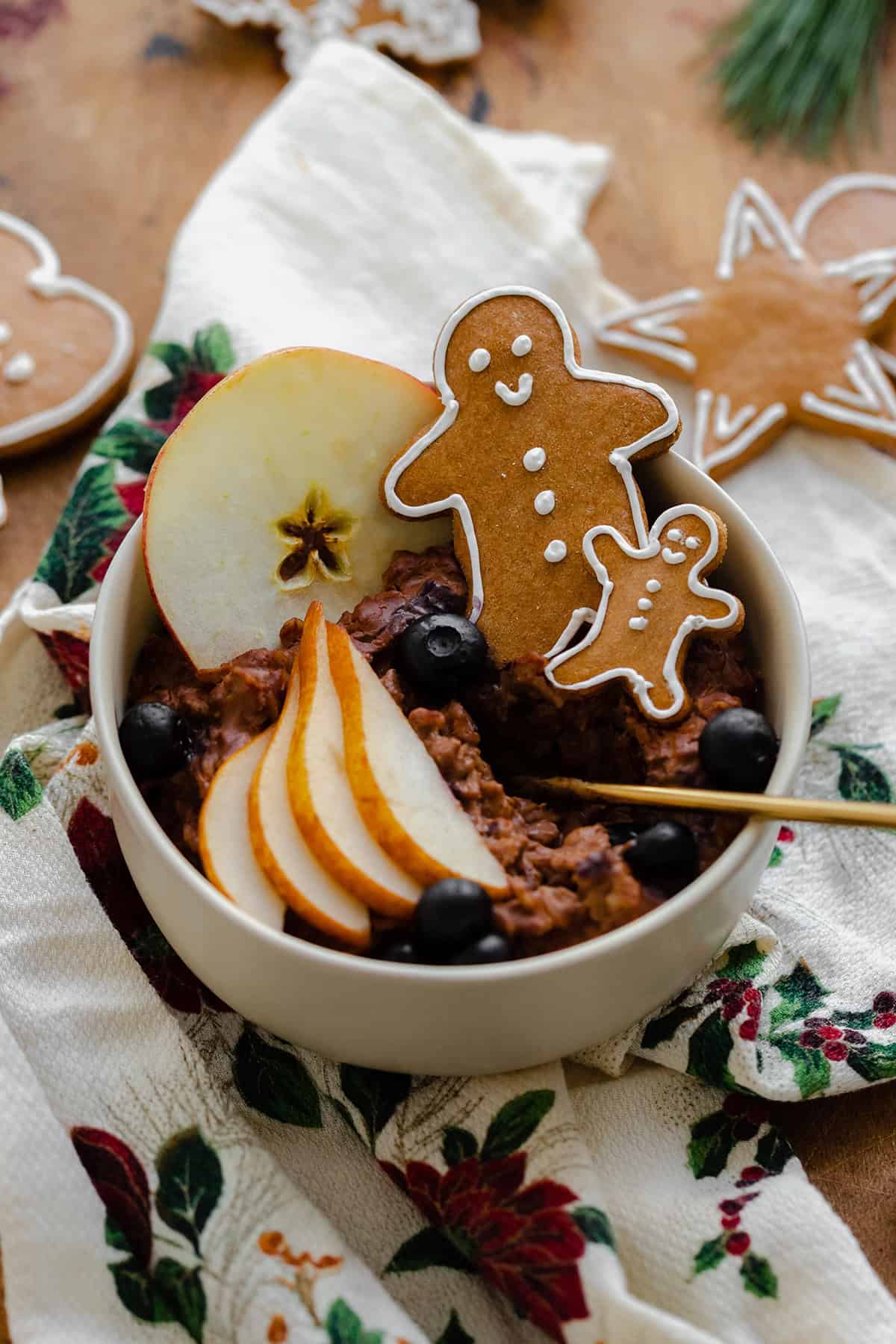 An overhead close up shot of Gingerbread oatmeal in a beige bowl garnished with sliced pear, a slice of apple, a few blueberries, and two gingrbread men. On light wooden background with a christmas tea towel under the bowl and gingerbread cookies around it.