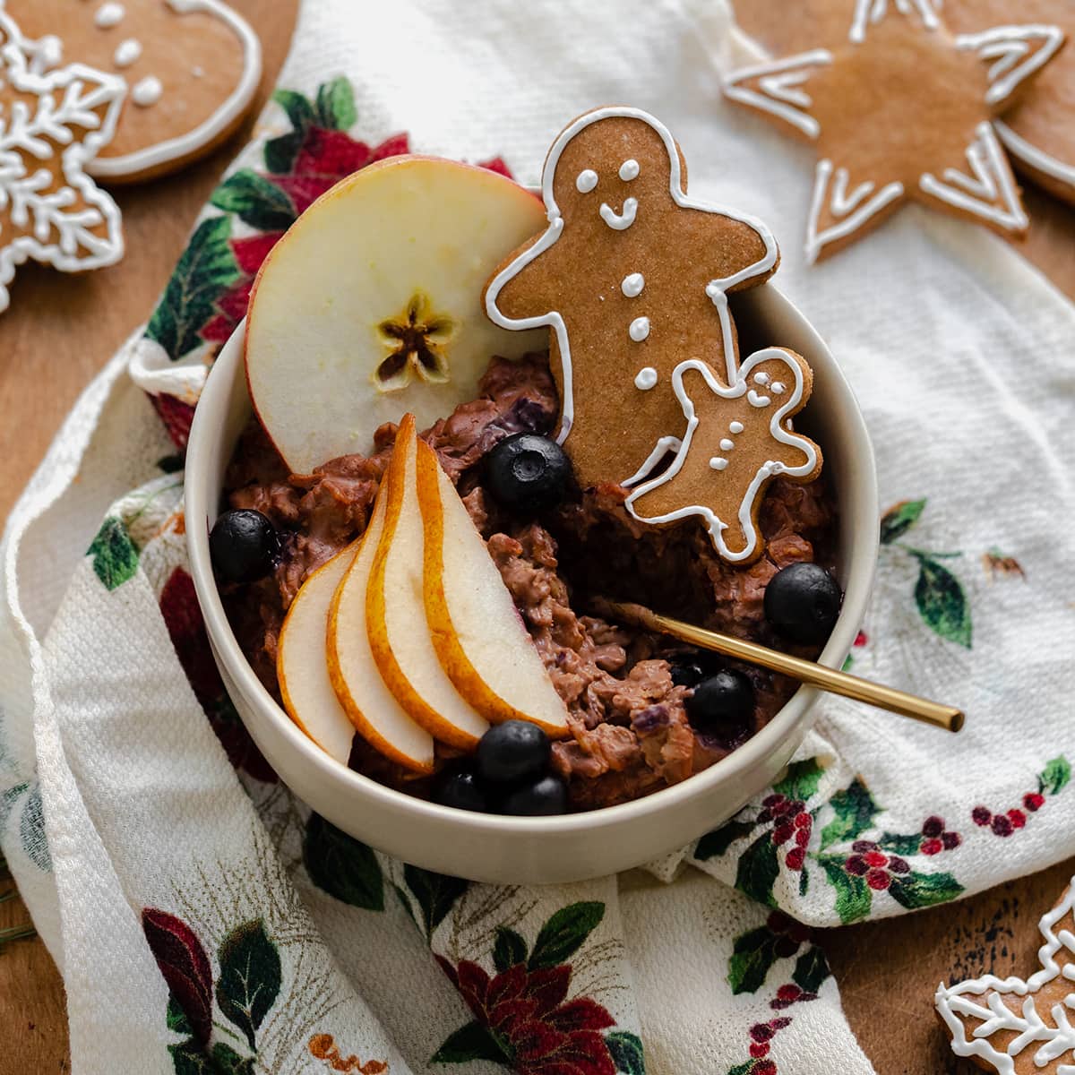 A square shot of Gingerbread oatmeal in a beige bowl garnished with sliced pear, a slice of apple, a few blueberries, and two gingrbread men. On light wooden background with a christmas tea towel under the bowl and gingerbread cookies around it.