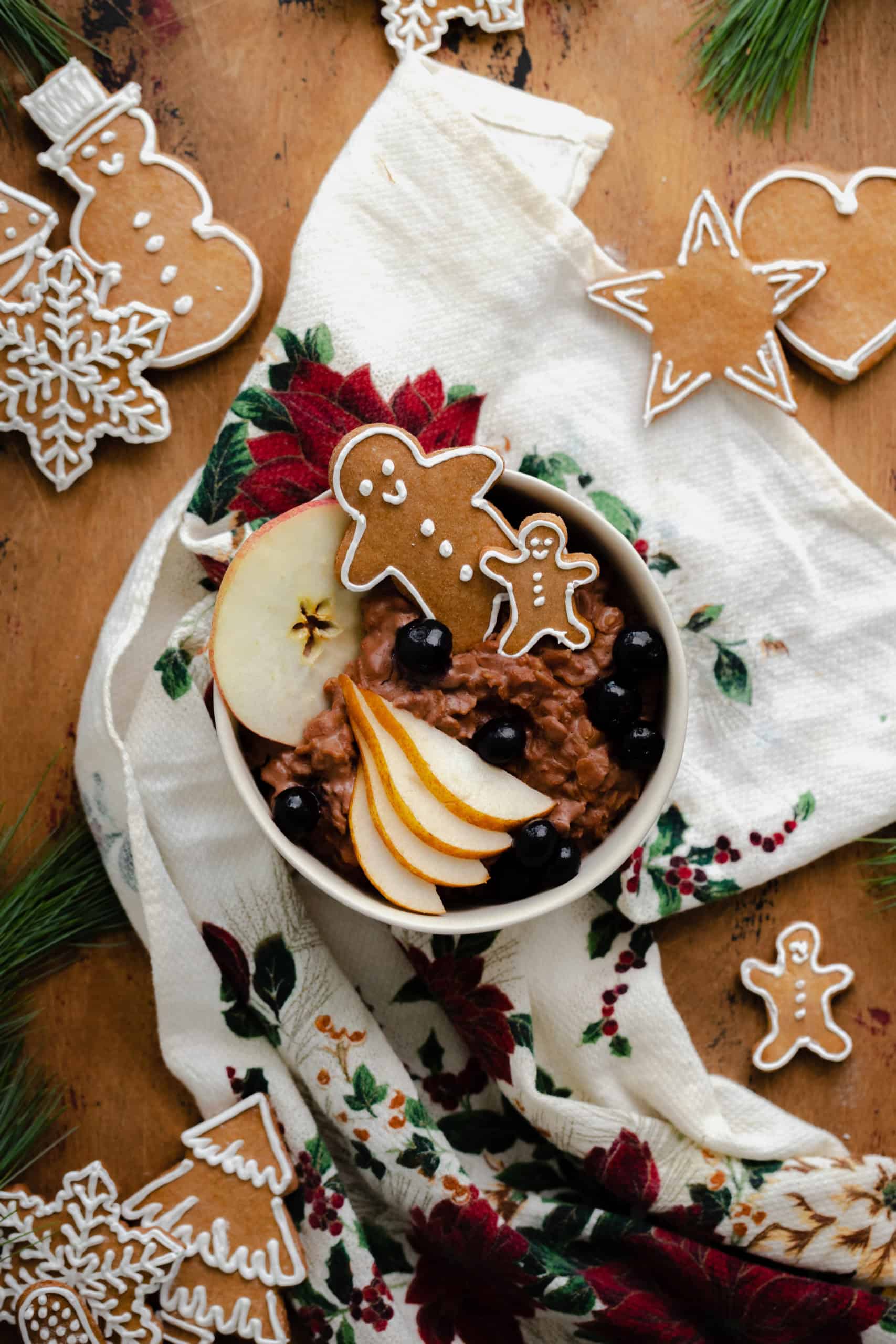 An overhead shot of Chocolate Gingerbread oatmeal in a beige bowl garnished with sliced pear, a slice of apple, a few blueberries, and two gingrbread men. On light wooden background with a christmas tea towel under the bowl and gingerbread cookies around it.