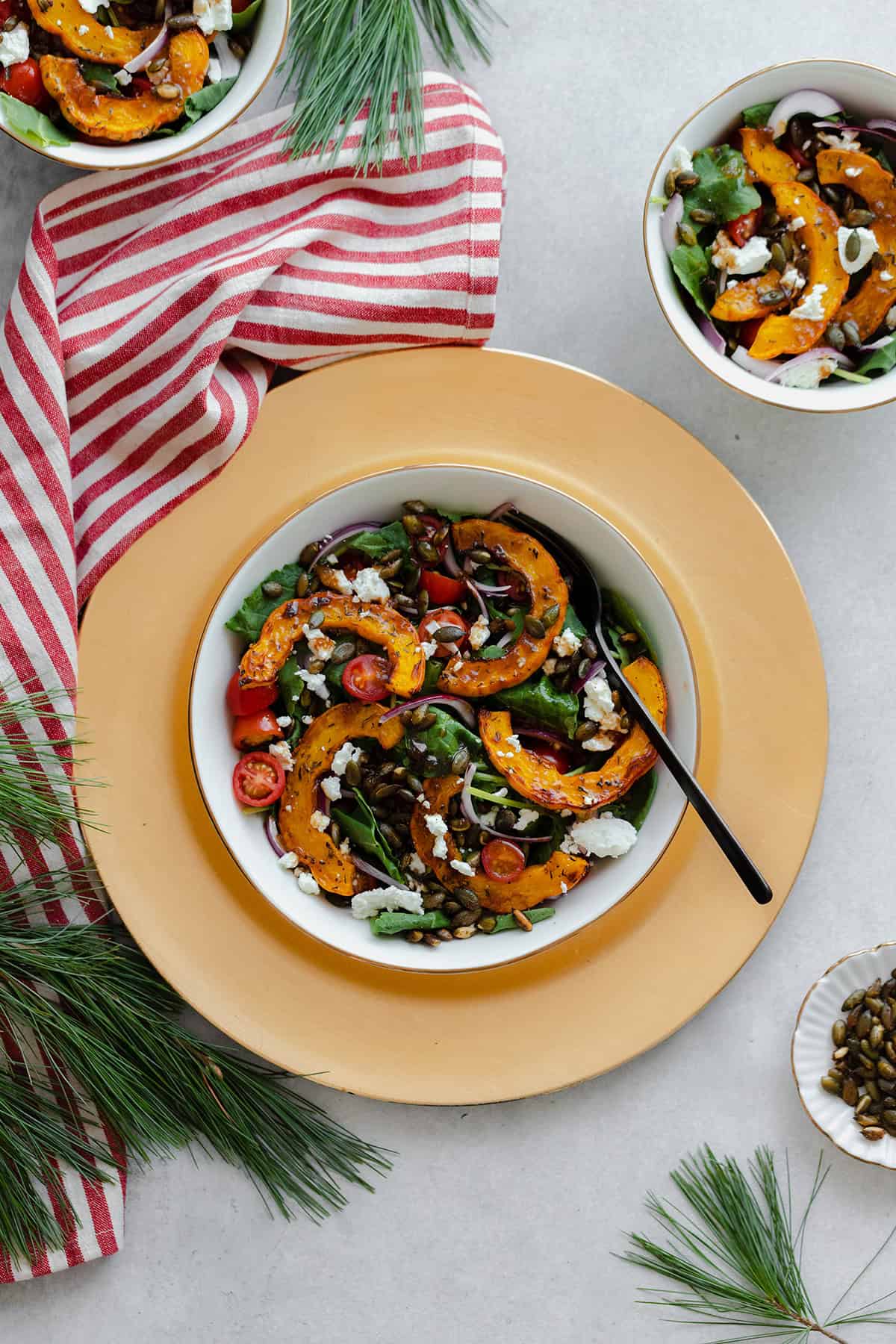 A photo of delicata squash salad in a white bowl with a black fork. Bowl on a decorative gold plate with a red and white tea towel on the left and a decorative some decorative pine branches.