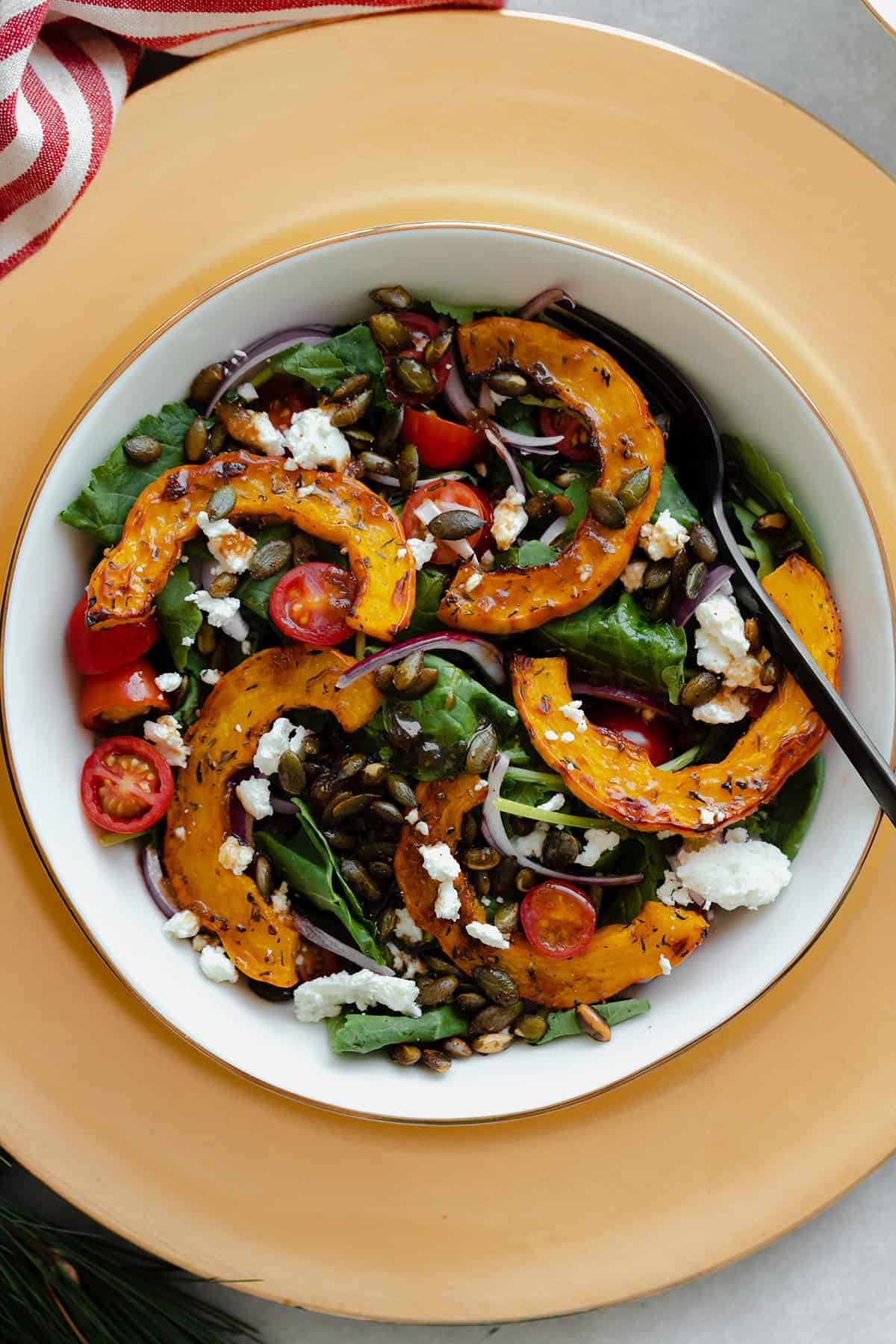 A close up of a salad with delicata squash, baby kale, cherry tomatoes, goat cheese, and red onion in a while bowl on a white decorative plate.