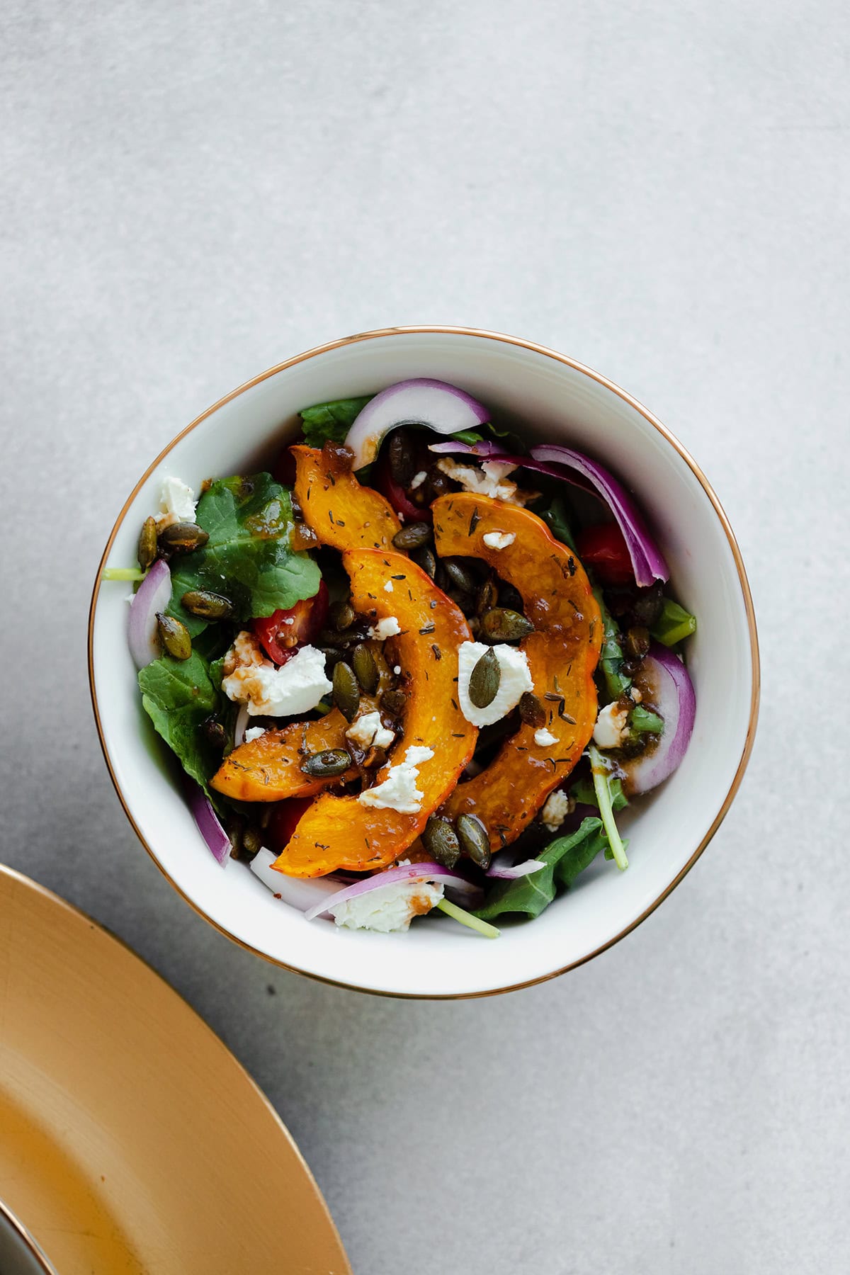 A small white bowl on a grey background with delicata squash kale salad, toasted pumpkin seeds, and crumbled goat cheese.