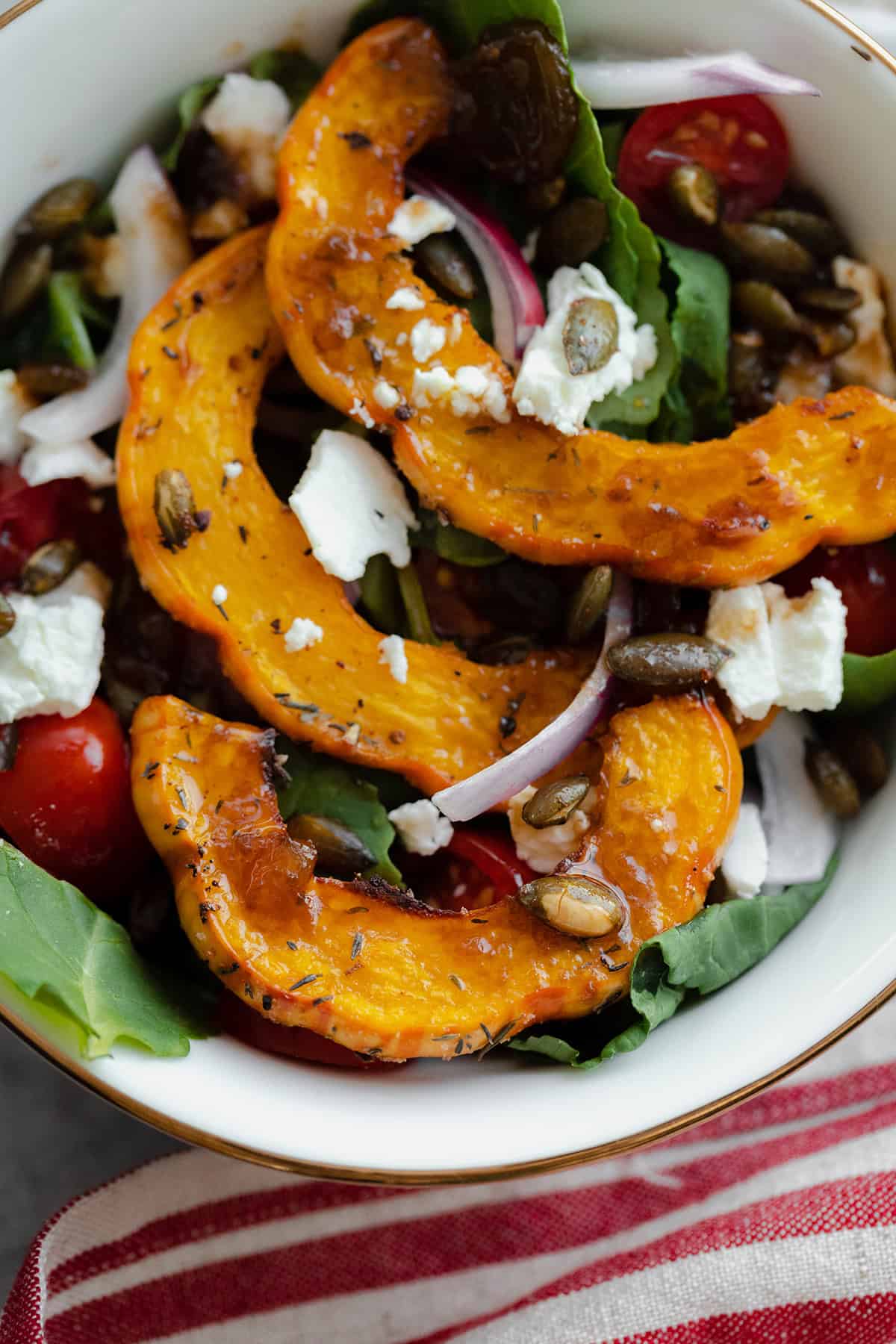 A close up of a small white bowl with a salad made with delicata squash, baby kale, goat cheese, tomatoes, and red onion.