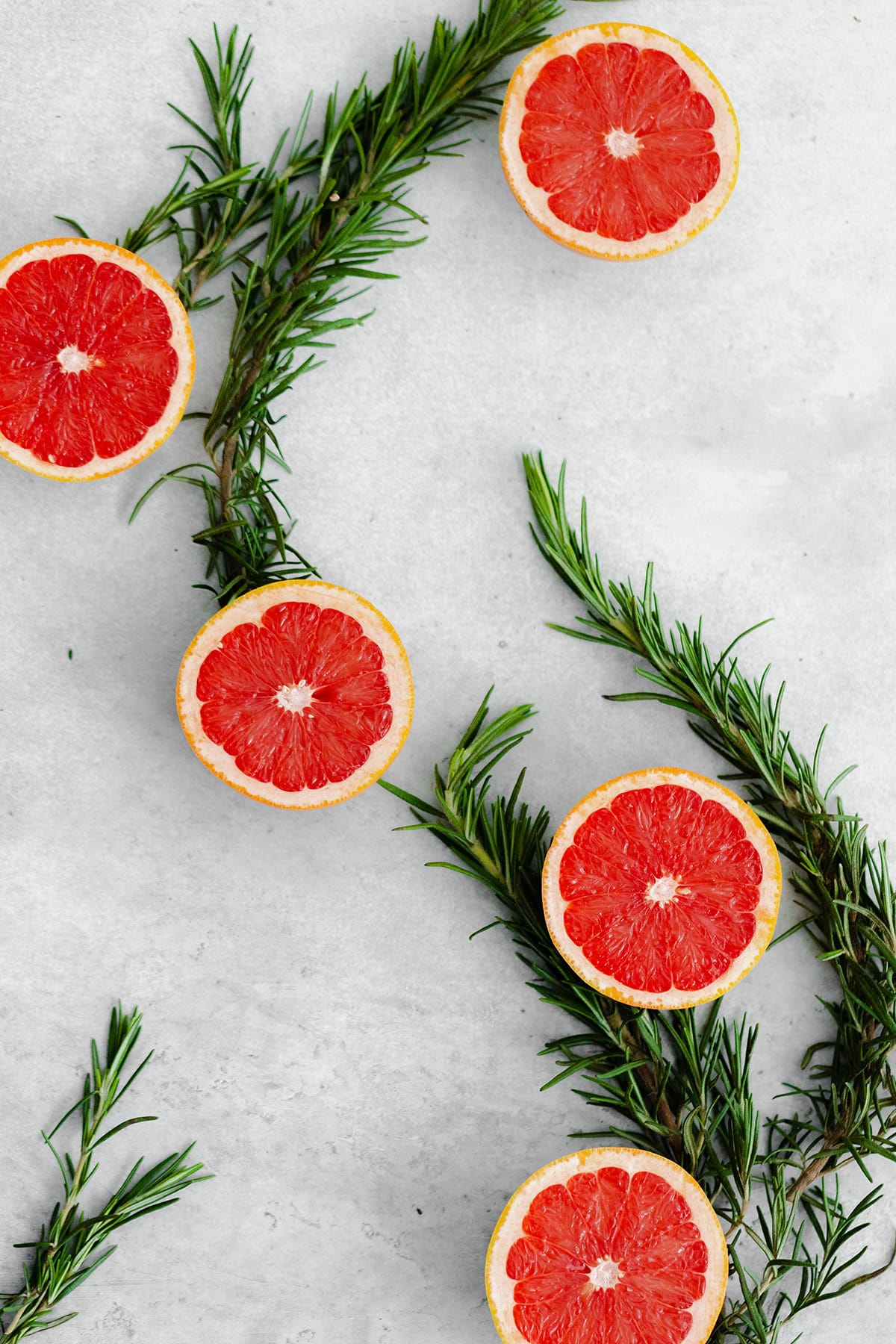 Grapefruits cut in half, laid out in an S shape with fresh rosemary connecting each piece. Cut side up on a gray background.