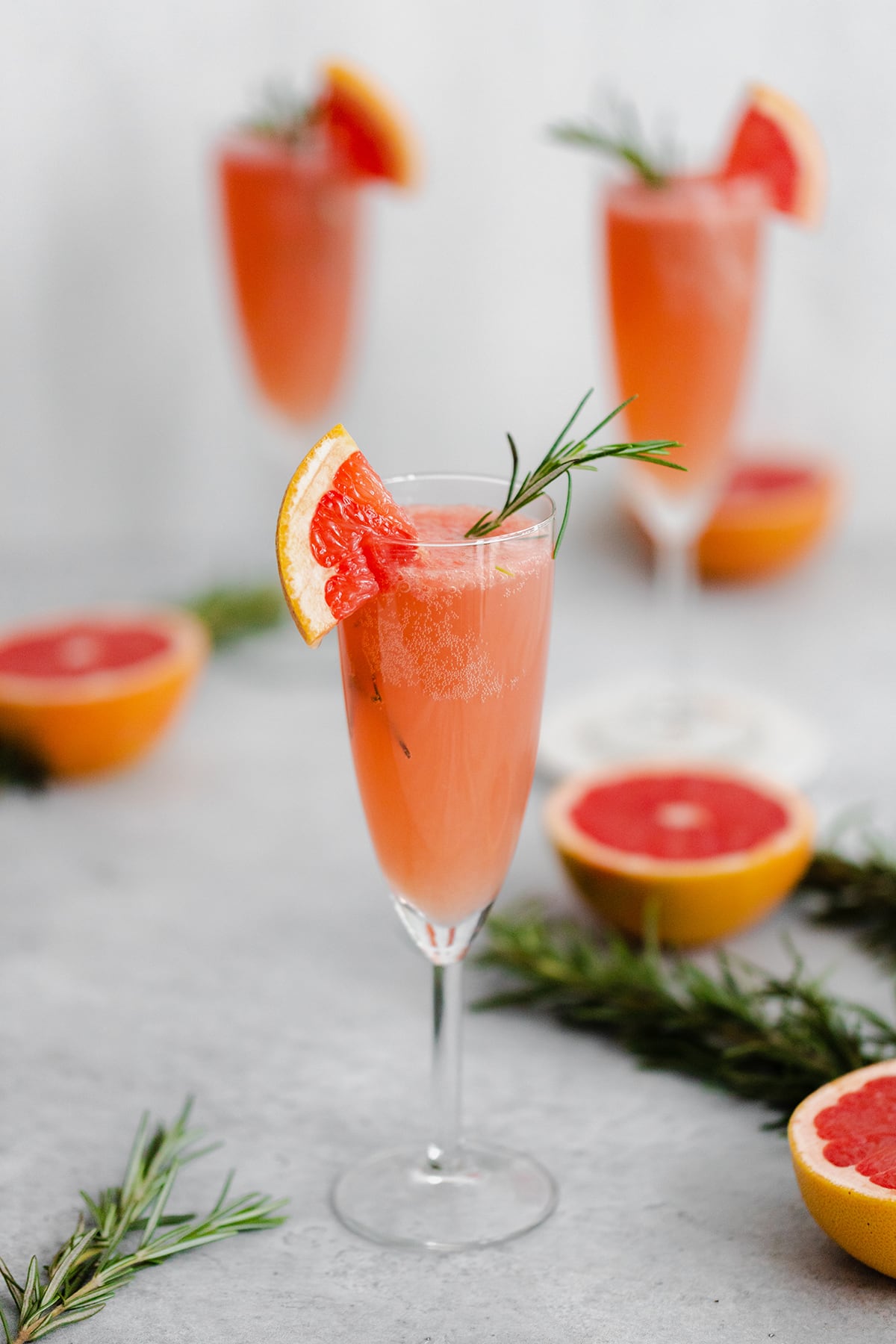 Grapefruit Mimosa in 3 flute glasses garnished with fresh rosemary and a quarter of a slice of grapefruit on the rim. Grapefruits as decoration in the background.